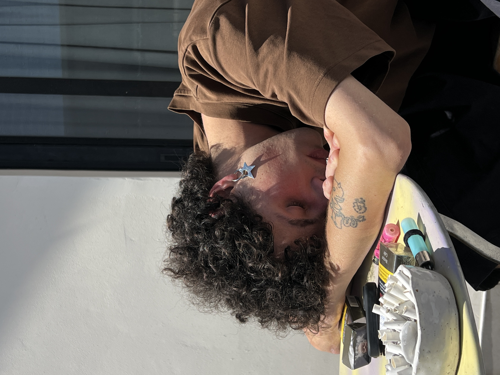 A young adult with curly dark hair is asleep on a table outdoors, their head resting on their crossed arms. They are wearing a brown t-shirt and have a star-shaped earring in their left ear. On the table, there is an ashtray filled with cigarette butts, several packs of cigarettes, and two lighters. The background shows a white wall and a dark-tinted window. The scene appears to be on a balcony or patio. There are no discernible elements in the image that specifically indicate the location is Tel-Aviv, Israel.