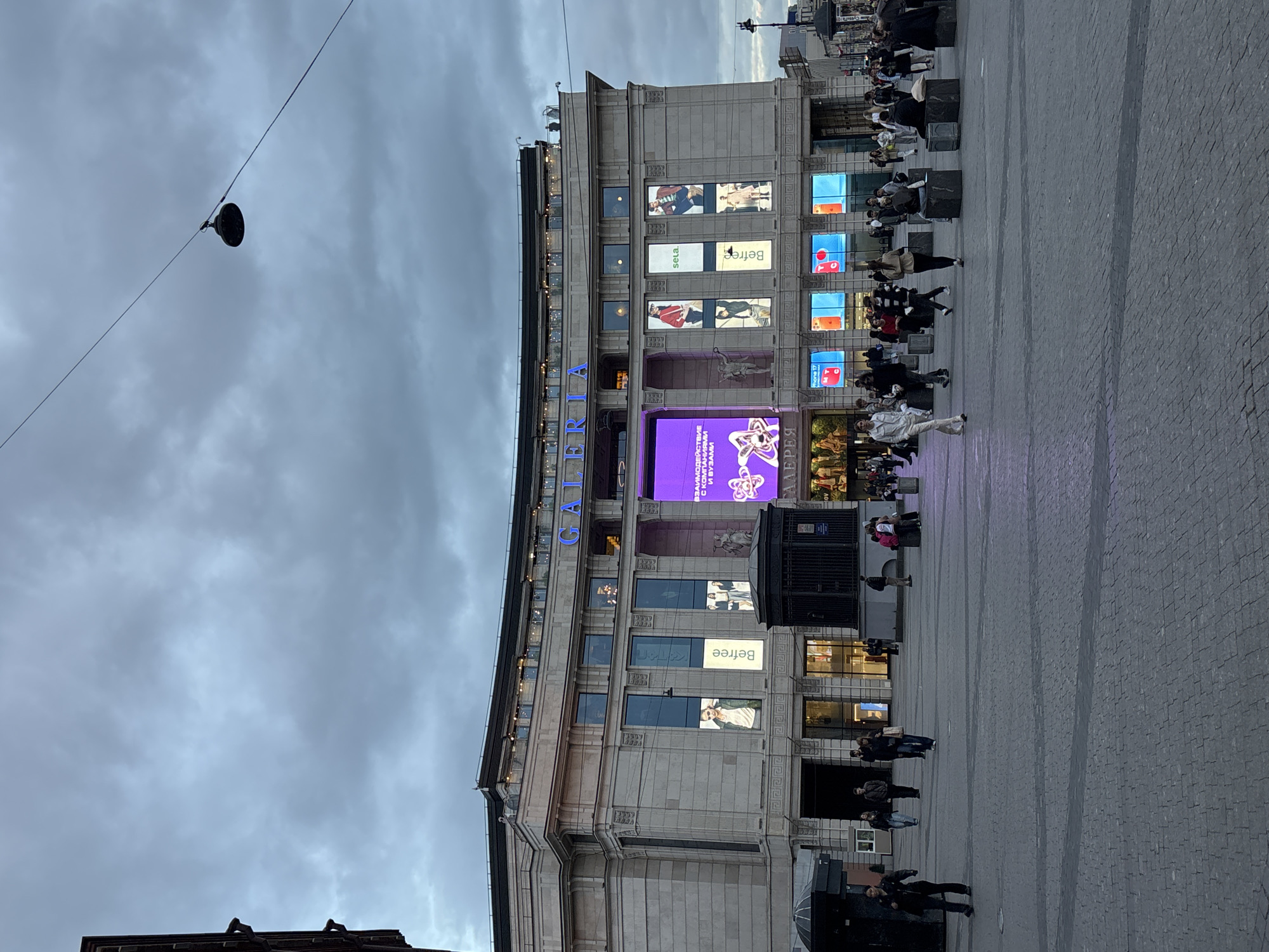 The image depicts the exterior of the "GALLERIA" shopping mall in Saint Petersburg, Russia. The building is multi-story, with a classical architectural style, featuring a gray facade. Large digital screens displaying advertisements are integrated into the building's design. People are visible on the sidewalk and in the vicinity of the mall. The sky is overcast. The foreground includes a paved area and the roofs of low-rise buildings. A street light on a wire is suspended overhead.