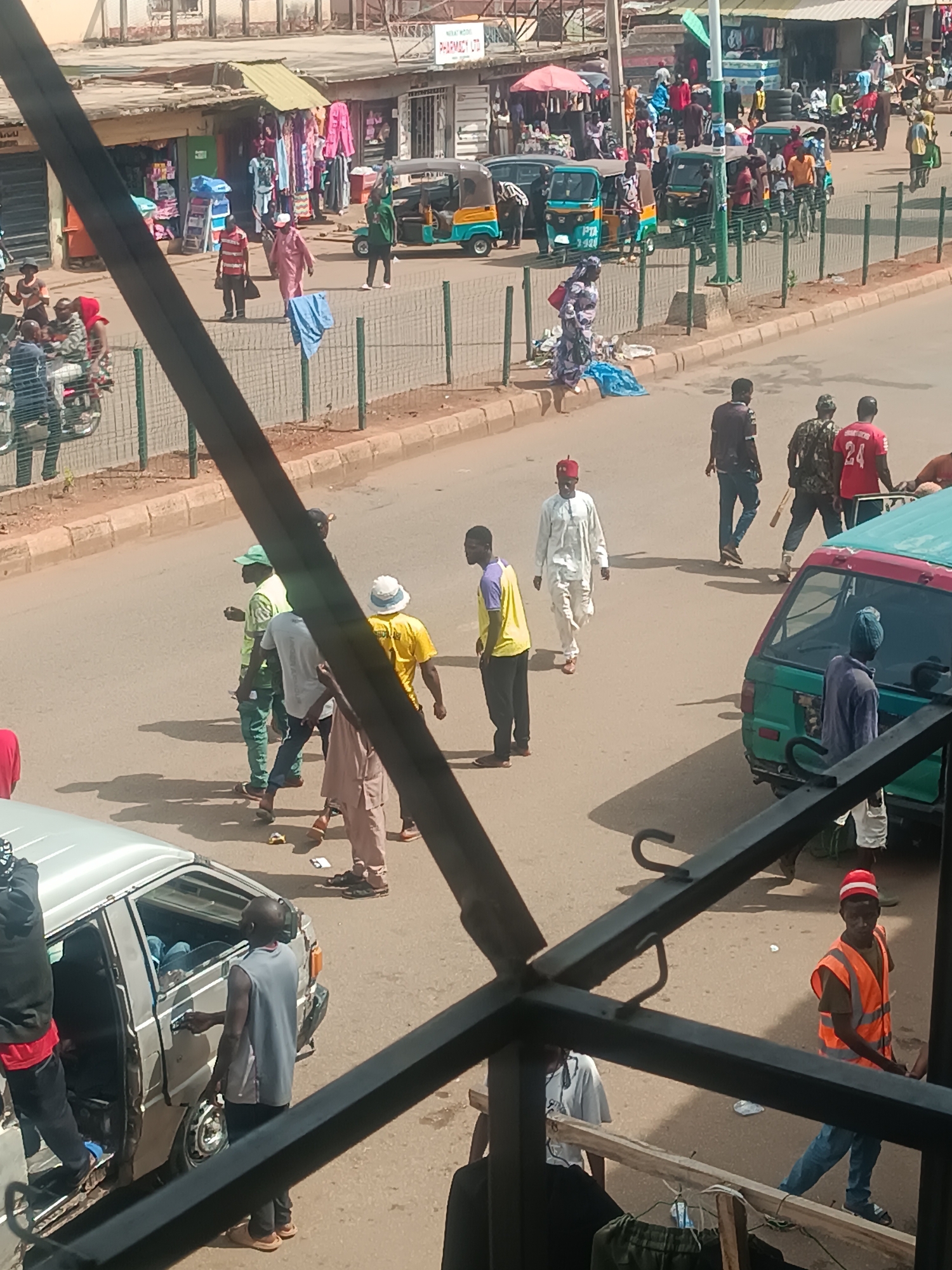 The image, taken from an elevated position, depicts a street scene in Bukuru, Nigeria. In the foreground, a dark metal balcony structure partially obscures the view. Below, a silver van has passengers entering and exiting. Several people walk along the street. A group of men in casual clothing is visible, as well as a man in traditional white attire and a red fez. Further away, there's a man wearing a red shirt with the number "24" on the back. Vehicles, including "Keke Napep" tricycles, are moving on the street, and shops line the sidewalk in the background.