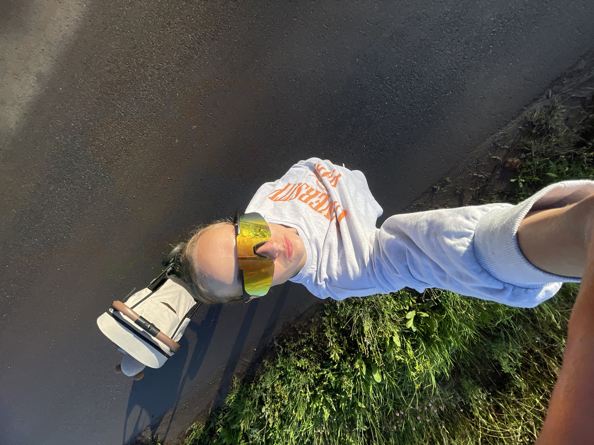 A young woman, wearing mirrored sports sunglasses and a white sweatshirt with orange lettering, is captured in a high-angle selfie. She is standing on an asphalt surface adjacent to a grassy area. To her left, a white stroller with a brown handle is visible. The scene appears to be outdoors, possibly a park or a walking path, under natural lighting. No specific landmarks identifying the location in the city of Подшивалово, Russia are present in the image.