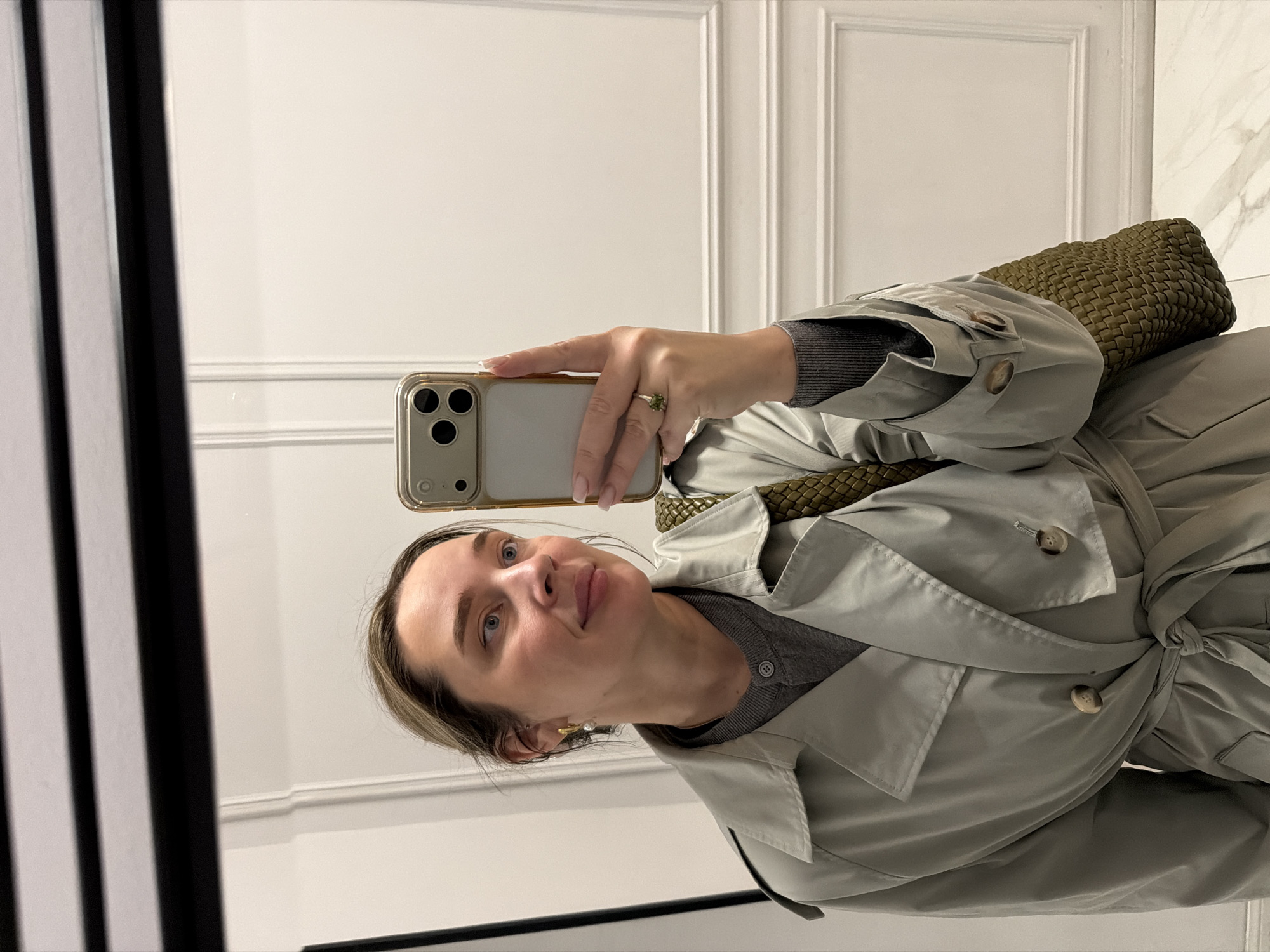 An eye-level mirror selfie captures a woman dressed in a light grey or sage-colored trench coat, worn over a dark grey knit top. She holds a gold-edged smartphone with a clear case in her right hand, taking the photograph. Her right hand is visible, wearing a prominent ring with a green, possibly raw-cut, gemstone on her index finger. Her light brown hair is styled back, and her face shows a calm, pleasant expression with a slight smile, highlighting her light blue eyes. A golden earring, possibly with a pearl or similar detail, is visible on her left ear. Over her right shoulder, a textured, dark olive-green woven bag is visible. The trench coat is tied at the waist and features visible buttons on the sleeves and lapel.
The background, reflected in the mirror, consists of a bright white wall adorned with classic decorative molding (boiserie panels). To the right, a hint of a light-colored marble wall can be seen. On the left side of the reflection, vertical black and white lines are visible, likely from the mirror's frame or an adjacent architectural element. The setting appears to be a well-lit indoor environment. The context places this scene in Moscow, Russia. No visible text is present in the image.