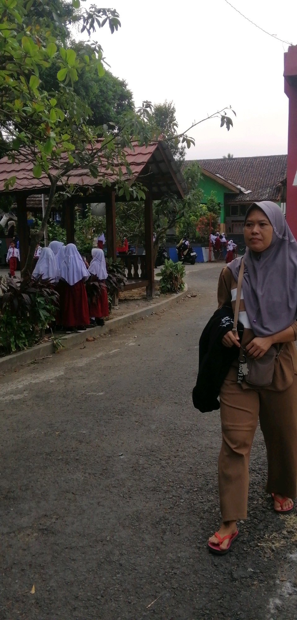 In Ajibarang, Indonesia, a woman wearing a grey hijab and brown uniform stands on a paved road. She has a light-colored shirt and pants, and is carrying a black jacket over her left arm, with a small brown crossbody bag hanging from her right shoulder. She is wearing pink flip-flops. Behind her, several school-aged children in white hijabs and red skirts, and one in a white and red uniform, are gathered under a wooden gazebo with a red tiled roof. The gazebo is surrounded by greenery and appears to be part of a school campus, with a building visible in the background. Several motorcycles are parked on the side of the road. The overall scene depicts an outdoor environment, likely a school, with a mix of adults and children.