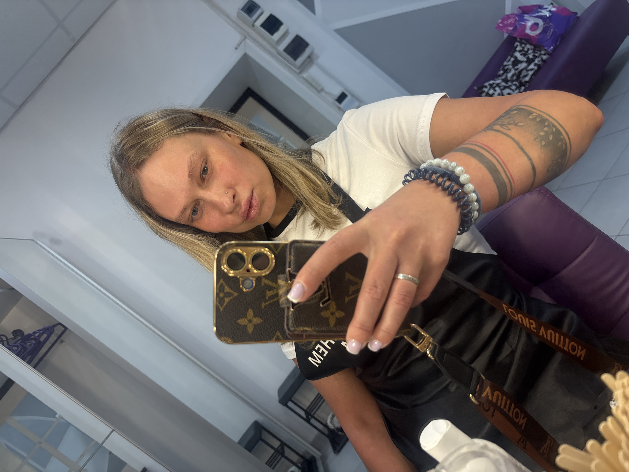 A fair-skinned woman with blonde hair takes a selfie indoors. She wears a white t-shirt and a black apron with brown straps. Her left arm, featuring a tattoo, is raised. A bracelet adorns her wrist. She holds a Louis Vuitton phone case in her hand. The background includes a purple chair, a cabinet, and a window. Based on metadata, the photo was taken in Perm, Russia.