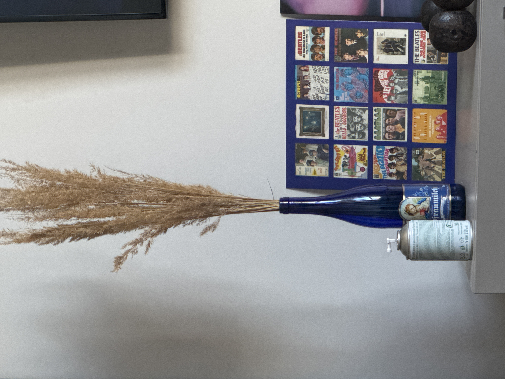 The image shows a still life composition against a light gray wall. A bundle of dried wheat stalks is placed in a dark blue glass bottle labeled "Fraumilch." Next to the bottle is a spray can, also placed horizontally. Behind the bottle, there is a framed collage featuring various album covers of The Beatles. Above the collage, there are a few dark-colored, spherical objects. A black object is partially visible in the upper left corner of the frame. The location is not possible to determine from the image.