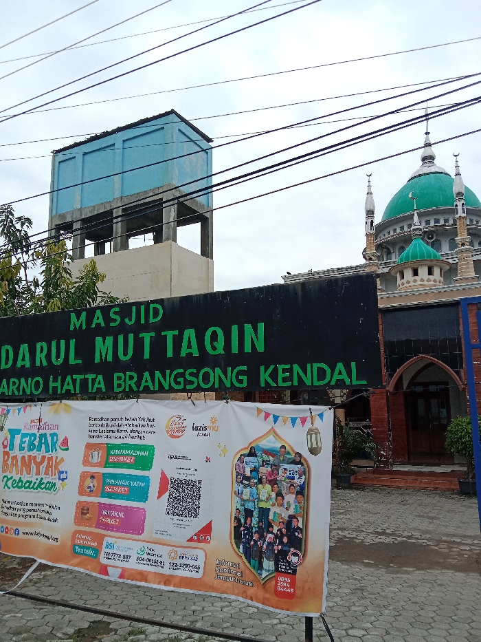 The image shows a mosque in the city of Kecamatan Brangsong, Indonesia, at the coordinates -6.948 latitude and 110.23 longitude. The mosque has a green dome and minarets, and is located in a residential area. The mosque is surrounded by a brick wall and a gate with a black sign that says "MASJID DARUL MUTTAQIN ARNO HATTA BRANGSONG KENDAL". There is a water tower to the left of the mosque, and a large banner in front of the mosque, with a QR code on it. The banner also says "Ramadhan penuh berkah, Yay Mu akan senantiasa hadir untuk berkontribusi, bersama Lazismu Kendal, dengan cara transfer atau iuran derma..." in Indonesian. The background is a cloudy sky. There are no people visible in the image.