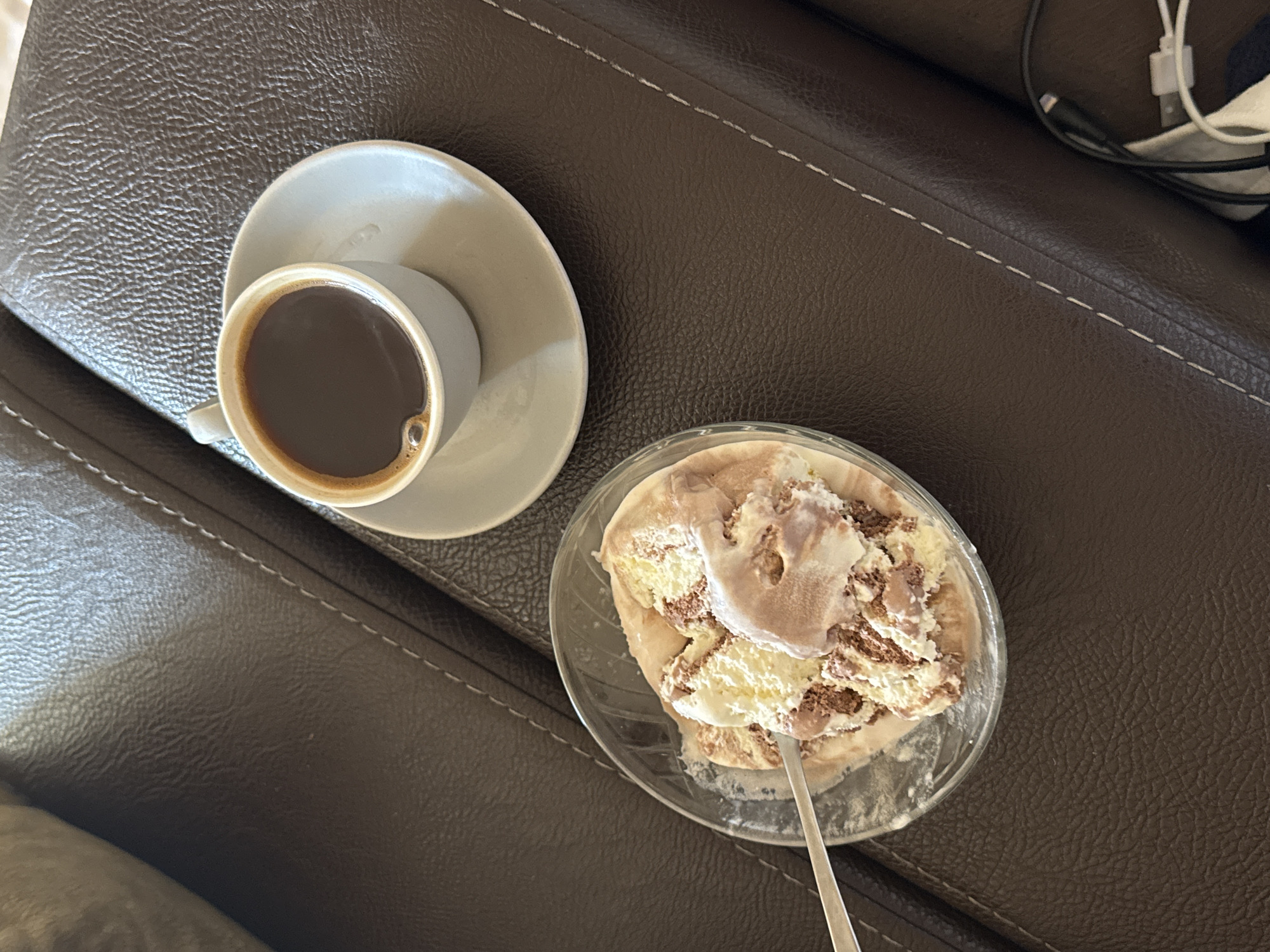 The image presents a top-down, slightly angled view of a moment of relaxation and indulgence in an indoor setting, likely in Yerevan, Armenia. A cup of coffee and a bowl of ice cream are placed on a dark brown, textured surface, which appears to be a sofa or armchair, distinguished by prominent white dashed stitching.
In the upper left portion of the frame, a light gray ceramic coffee cup with a matching saucer holds a dark liquid, presumably coffee, with a small bubble visible on its surface.
Towards the lower right, a clear glass bowl contains several scoops of ice cream. The ice cream exhibits a marbled pattern, combining creamy white or vanilla portions with rich brown or chocolate swirls, suggesting a multi-flavored dessert. A silver-colored spoon is partially submerged in the softened ice cream.
The background reveals more of the dark brown, textured upholstery. In the top right corner, a glimpse of white and black cables, possibly belonging to a charger or other electronic devices, can be seen, indicating a contemporary living space. The scene evokes a sense of comfort and pleasure, combining the warmth of coffee with the coolness of ice cream. There is no visible text in the image.