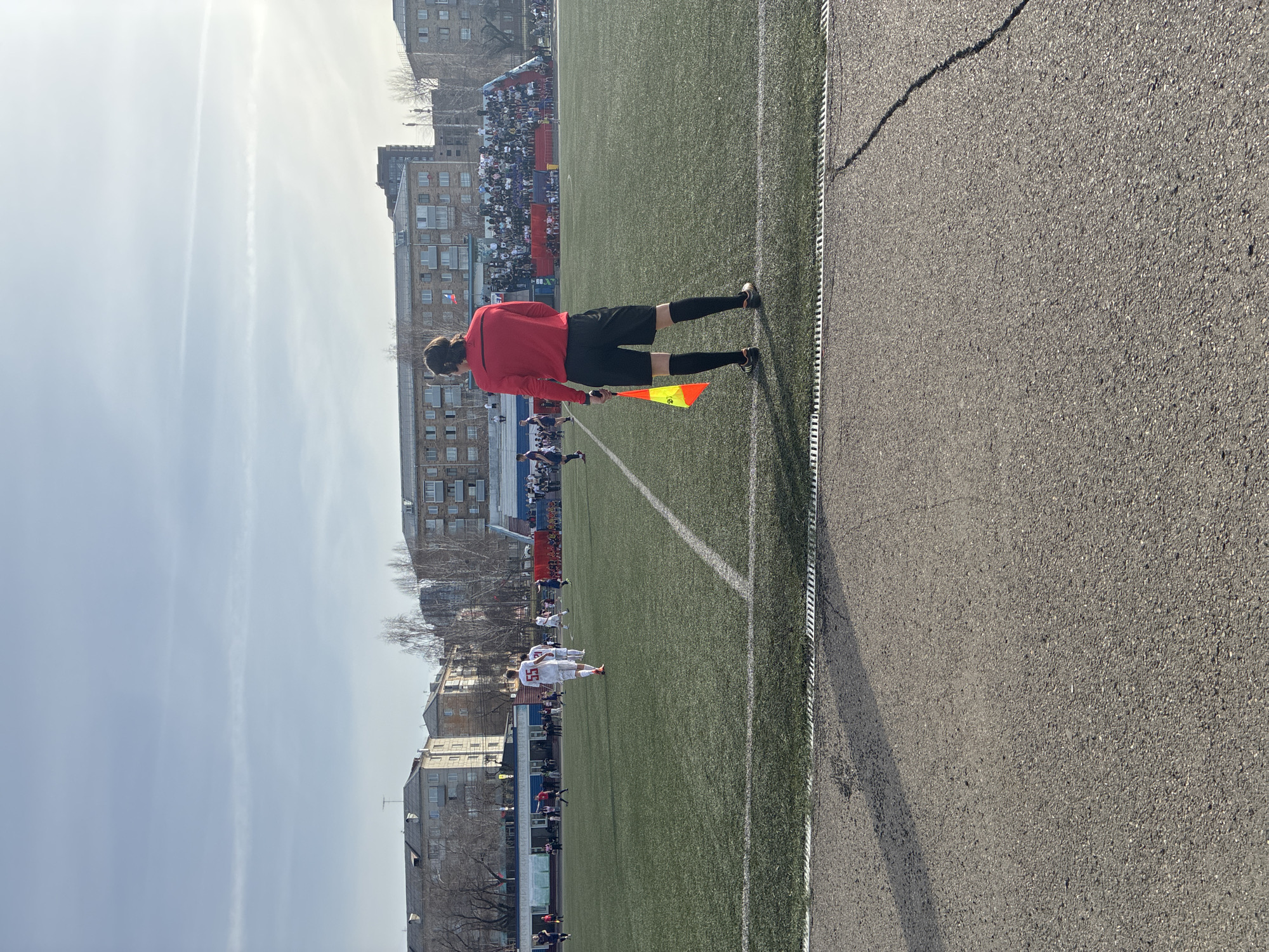 The image, rotated 90 degrees clockwise, captures an outdoor football (soccer) match from a side-line perspective. In the foreground, an assistant referee (linesman) is observed from behind, dressed in a red long-sleeved shirt, black shorts, and black socks, holding a yellow and orange flag. Their shadow is cast on the asphalt path to their right, indicating bright sunlight.
To the left of the linesman, the artificial turf pitch is visible, marked with white lines. Several players are on the field, including one prominently featuring the number "55" on the back of their white jersey. Other players in both white and darker kits are scattered across the pitch. Behind the field, spectators are gathered in stands, appearing as a dense crowd.
The background consists of several multi-story buildings, likely residential or commercial, under a bright, slightly hazy sky with subtle contrails. Bare trees, suggesting a season other than summer (e.g., late autumn, winter, or early spring), are visible among the structures. A small flag with white, blue, and red horizontal stripes (resembling the Russian flag) is affixed to one of the distant buildings. The scene conveys a daytime event under sunny weather at an urban sports complex. No specific emotions are discernible.