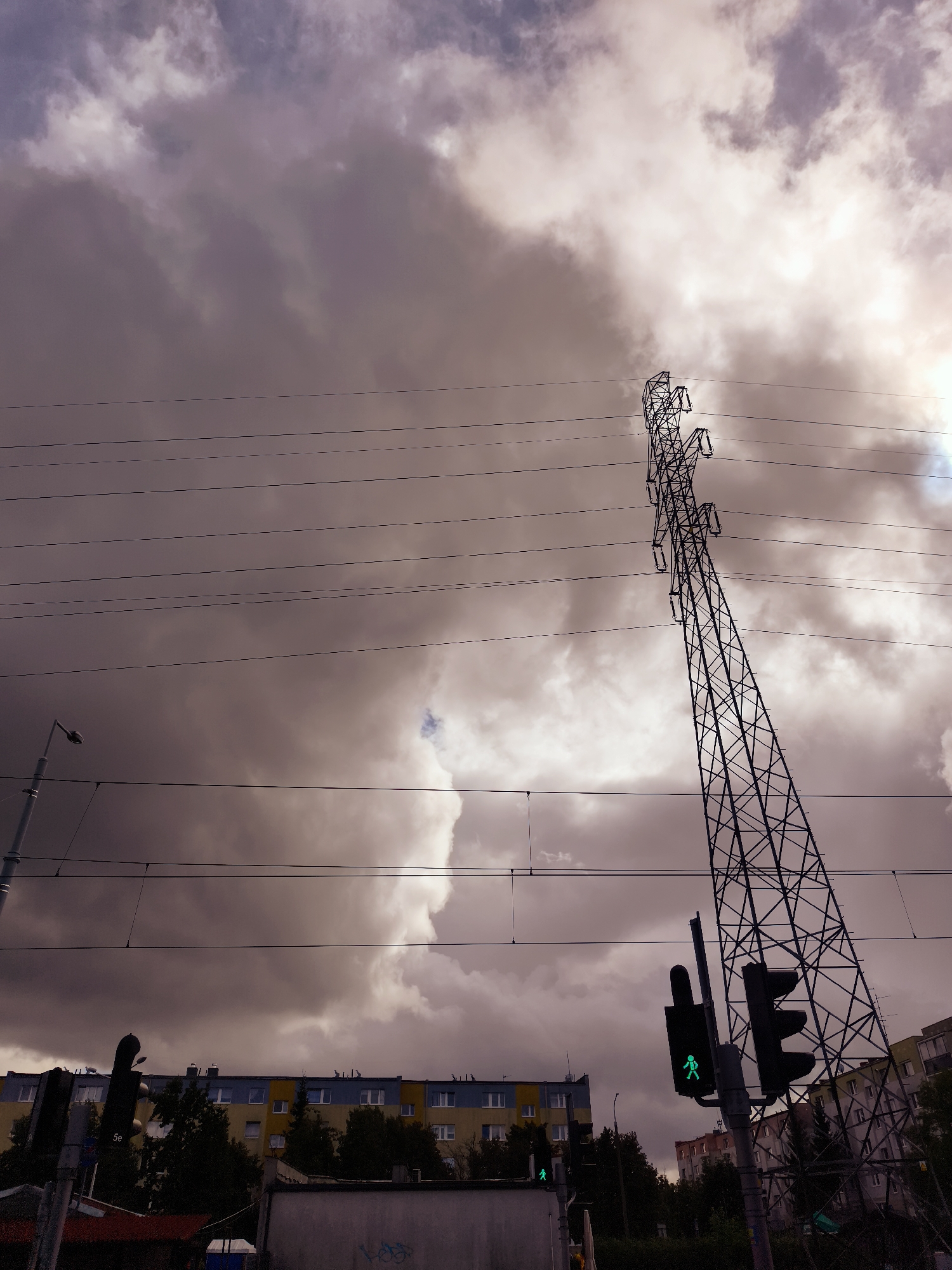 This is a medium-shot, low-angle, vertical photograph of an urban scene dominated by a massive, latticed metal electricity pylon. The pylon, a prominent feature, is tilted and extends diagonally upwards from the lower right towards the upper right corner. Multiple power lines stretch horizontally across the frame, originating from or passing by the pylon. The sky is filled with dramatic, heavy, and partially illuminated storm clouds, casting a dark, moody atmosphere over the scene.
In the background, a row of apartment buildings stretches horizontally across the mid-ground. These buildings are distinct with alternating sections of yellow and blue facades, punctuated by numerous windows. Below the apartment buildings, dense green trees are visible.
In the foreground and mid-ground, there are elements of urban infrastructure. A traffic light, positioned on the right side of the frame, displays a green pedestrian signal, showing a walking figure. To the left, another traffic light, partially obscured, also appears to show a green pedestrian signal. A street lamp is visible on the far left side. The lower portion of the image reveals the top of a building with a red roof element on the far left, and a grey concrete structure with graffiti tags on the right.
The scene appears to be located in the city of Gdańsk, Poland. The overall impression is one of a city under a dramatic sky, with prominent industrial infrastructure and residential buildings. There are no visible people in the immediate foreground or engaging in any specific actions. The focus is on the architectural and atmospheric elements.