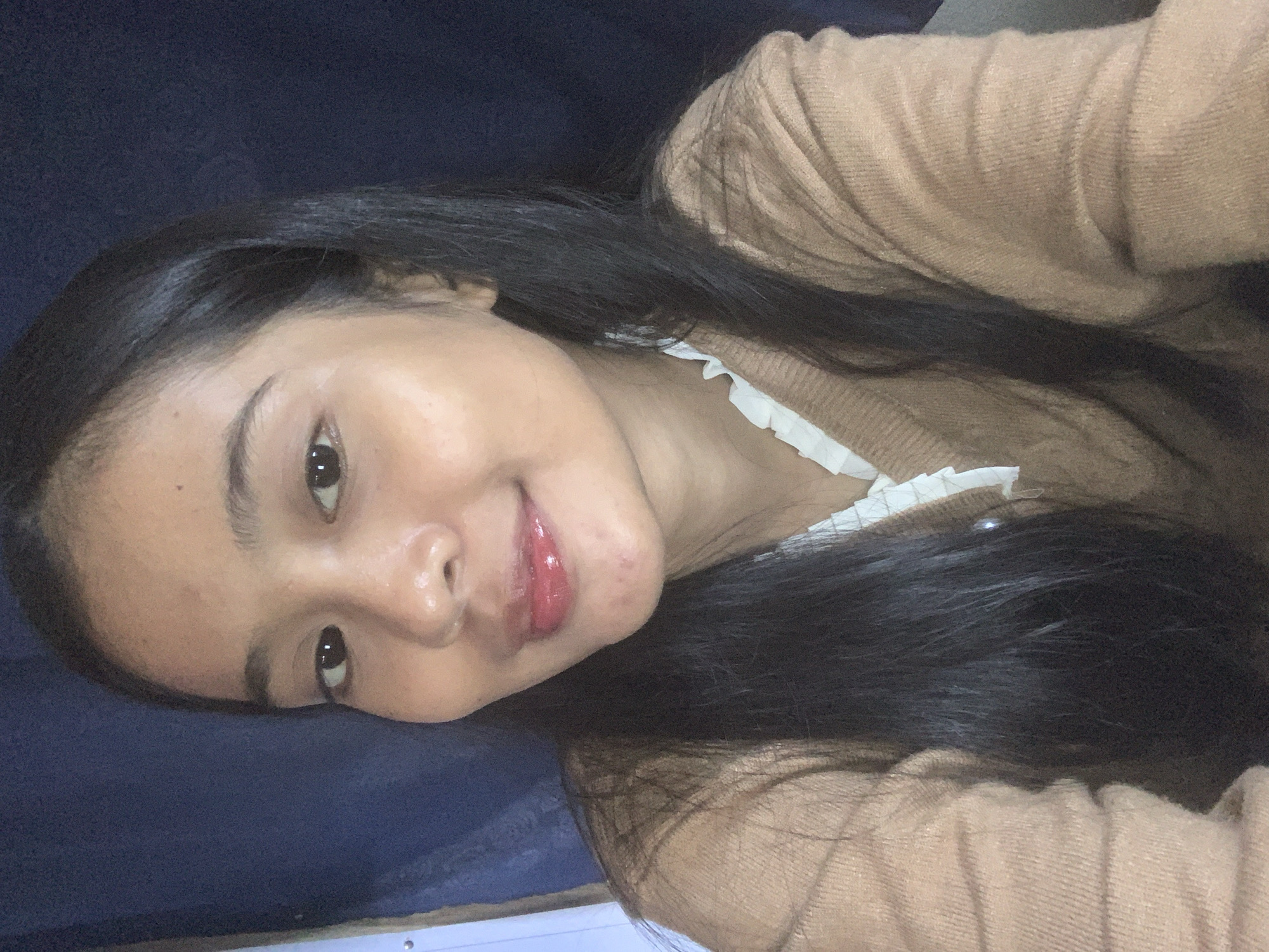 The image is a close-up selfie of a young woman with dark hair, facing the camera. She is wearing a tan cable-knit sweater over a white ruffled-collar top. Her lips are glossy and appear to have red lipstick. The background is a dark blue fabric. The image does not provide any information about the location in Sungai Meriam, Indonesia.