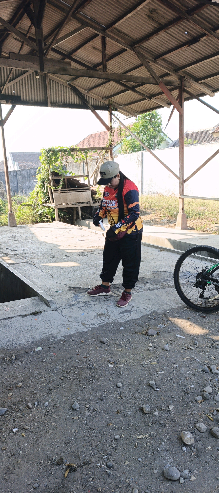 A person wearing a baseball cap, a long-sleeved multi-colored shirt with "RACING" text, black pants, and maroon sneakers stands outdoors under a corrugated metal roof supported by wooden beams. The person is looking down at their hands, which are holding a white object. To the right of the person, a bicycle wheel is visible. The ground is rough and made of dirt and gravel. In the background, there is a wooden structure with overgrown vegetation, houses with red roofs, and a white wall. The scene appears to be in an outdoor, possibly semi-abandoned, area within the city of Kebonan, Indonesia.