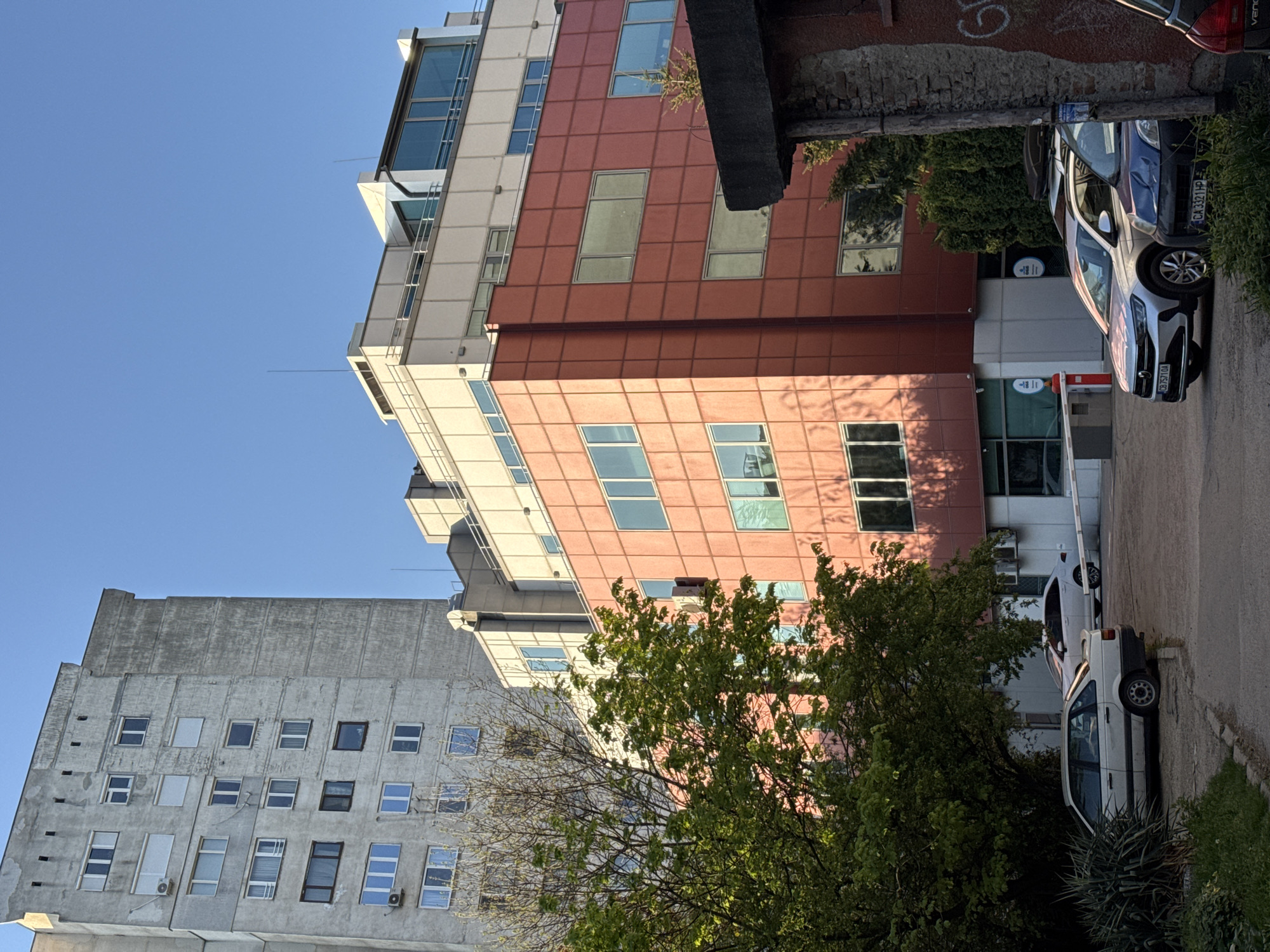 This street-level photograph, taken in Sofia, Bulgaria, captures an urban scene featuring two distinct buildings, parked cars, and trees under a clear, bright blue sky. The image is oriented vertically, rotated 90 degrees clockwise from its natural upright position.
In the foreground and midground, a modern building with a reddish-brown, grid-patterned facade and numerous large square windows dominates the right side of the frame. Its upper floors and architectural accents are a contrasting white or light grey. A small, round blue sign is visible near an entrance, next to a white barrier gate with a red arm, suggesting controlled access to a parking area or entrance.
To the left and further back, a taller, older building made of grey concrete is visible. Its facade is weathered, featuring numerous smaller, varied windows, some with air conditioning units beneath them. The building's roofline appears stepped.
Several vehicles are parked on an asphalt driveway on the right. A white SUV, possibly a Volkswagen T-Roc, is prominent, displaying the license plate "CB 7571 OA". Beside it, a blue-grey car with the license plate "CA 323 HP" is parked. Another older white car is partially visible further down the driveway. A rough brick wall with indistinct graffiti provides a backdrop near the blue-grey car.
Lush green trees with dense foliage are scattered throughout the midground, with some leafless branches also present, casting distinct shadows that suggest bright daylight, likely late morning or afternoon. The sky is uniformly clear and blue. The scene is quiet, with no visible human activity.