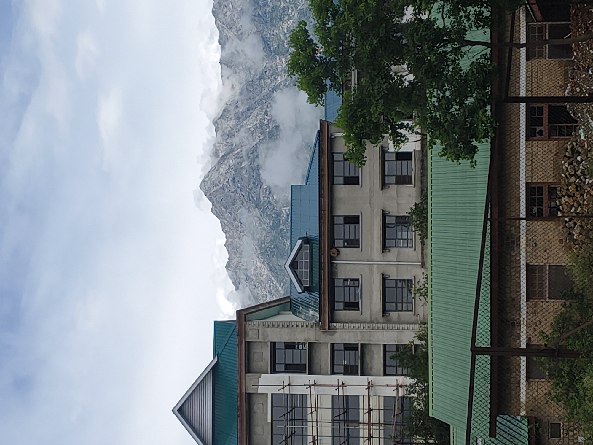 The image shows a multi-story building with a green metal roof, partially under construction with scaffolding visible on one side. The building is made of concrete and has numerous dark-framed windows. In the background, snow-capped mountains rise above the clouds, suggesting a high-altitude location. The foreground features another structure with a green metal roof, constructed with stone and brick walls, also appearing somewhat unfinished or in disrepair, with piles of debris and logs visible. A large green tree with dense foliage partially obscures the view of the buildings and mountains. The sky is overcast with scattered clouds. This scene is consistent with the landscape and architecture found in Dharamshala, India, known for its hilly terrain and the presence of the Dalai Lama's residence.