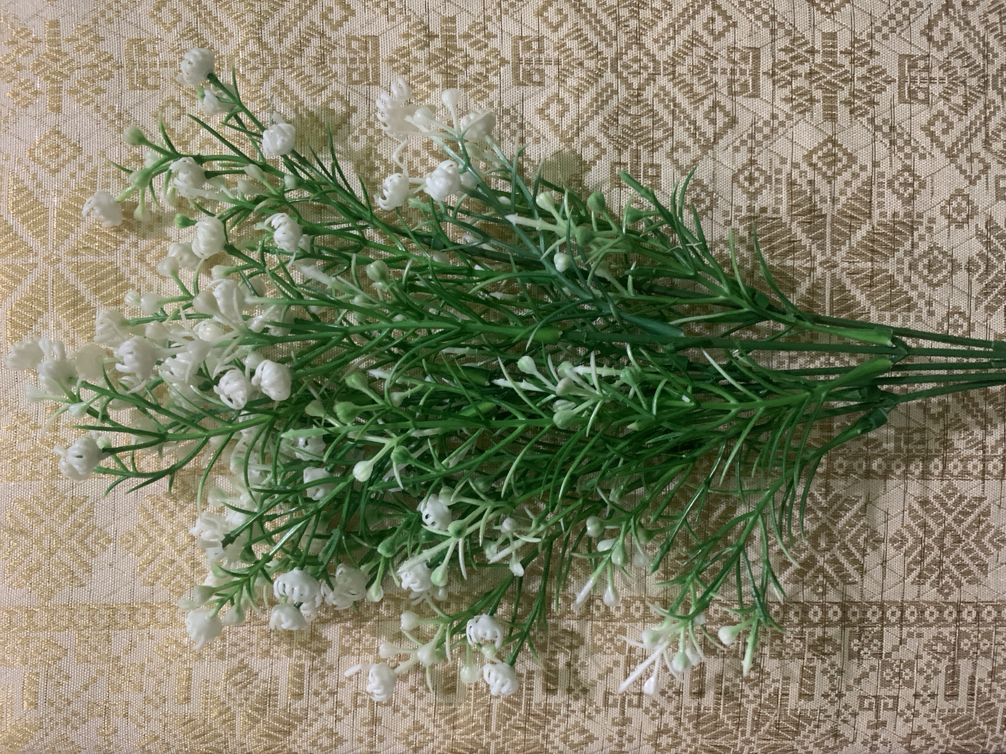 A close-up, top-down shot captures a bouquet of artificial white baby's breath flowers and green stems. The bouquet is positioned on a beige textile with a repeating geometric pattern in a darker brown or gold hue. The textile has a textured weave. The lighting suggests an indoor setting, likely a home or studio. The image does not contain any information to determine its location in Kuala Pembuang, Indonesia.