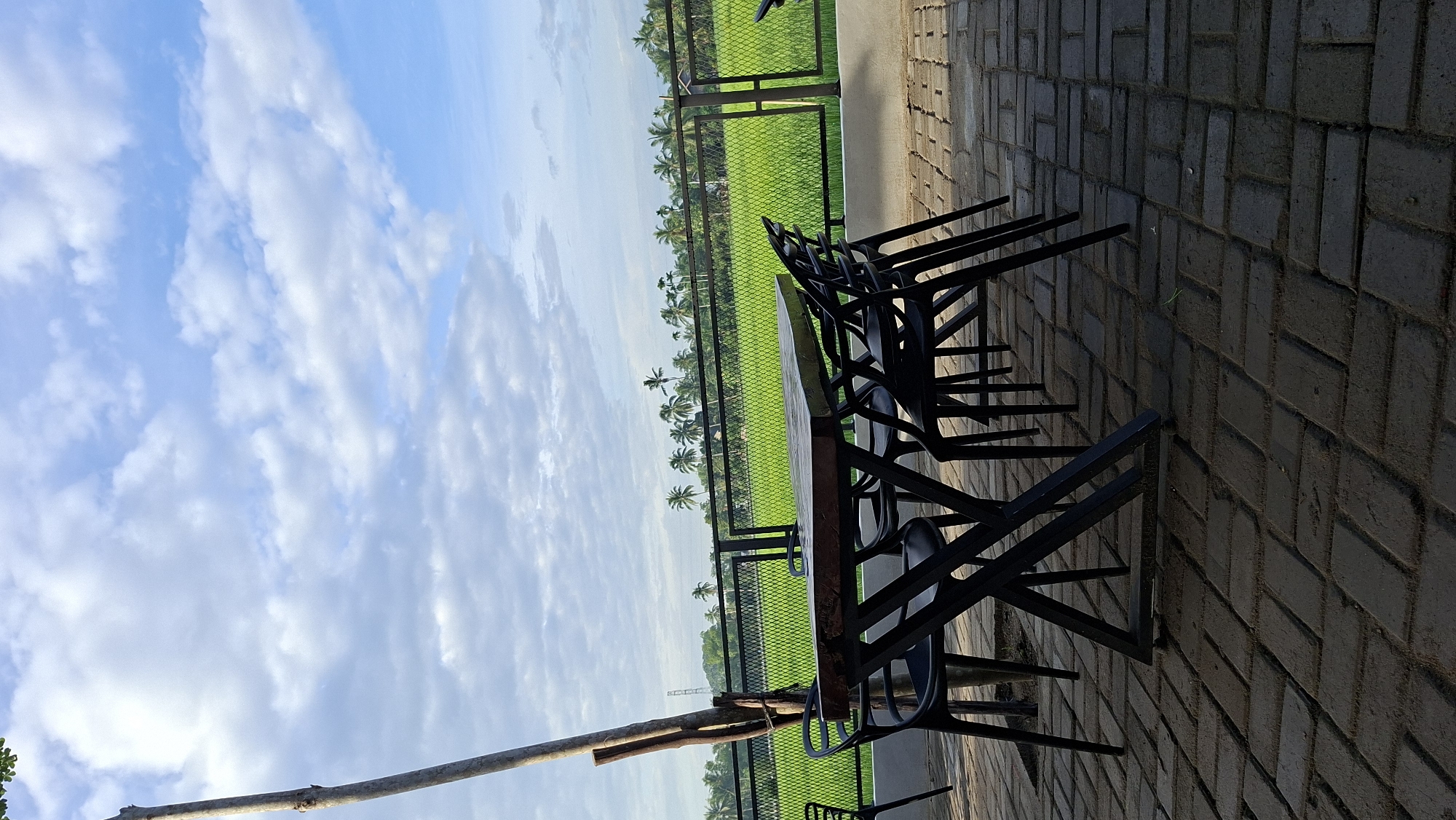 A wooden table with black chairs is set on a brick patio. Behind the patio, a green rice field stretches to the horizon, bordered by a fence and a line of palm trees. The sky above is blue with scattered white clouds. The scene is peaceful and rural, with no people present. The location is Keudebayu, Indonesia.