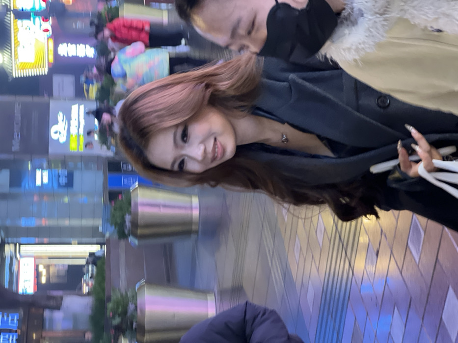 The image depicts a nighttime urban street scene in Yuzhong District, China. The primary subject, an East Asian female, appears in the foreground, slightly left of center. She has long, wavy, reddish-brown hair, is wearing a dark, possibly charcoal-colored, long coat over a dark top with a visible small pendant necklace, and displays a slight smile while looking towards the viewer's left. Her left hand holds several white shopping bags, and her fingernails are long and metallic-silver in appearance.
To her immediate right, a second East Asian individual is partially visible, mostly from the right side of their face, wearing a black face mask and a light-colored, possibly cream or beige, jacket with a fur-trimmed collar. This individual is oriented away from the camera. On the far left, the shoulder and arm of a third person, wearing a dark blue or black puffer jacket, are visible.
The environment is a brightly illuminated commercial street. The pavement consists of light-colored tiles with darker rectangular patterns and tactile strips. Two large, tapered, metallic-gold planters containing green foliage are positioned on the pavement. The background features multiple illuminated building facades and neon signs displaying various Chinese characters and some English text, including "Mercure." Several blurred figures, including one in a multi-colored puffer jacket and another in a red jacket, are visible in the distant background, indicating pedestrian activity. The scene is lit by artificial light sources from the surrounding buildings and street.