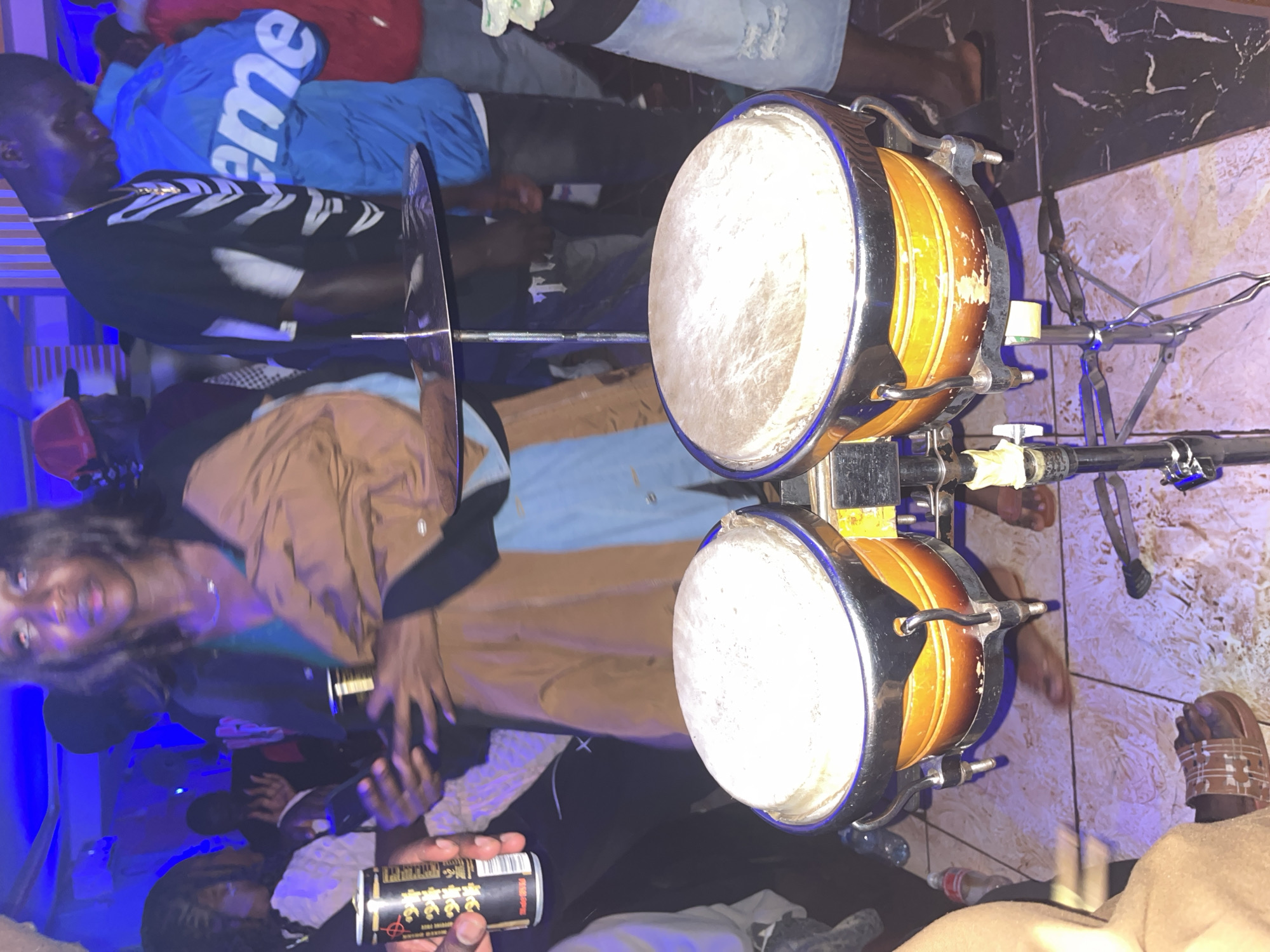 A low-angle, vertical shot captures a vibrant scene in what is identified as the city of Wumat, Nigeria. In the foreground, a pair of bongos rests on a stand, their surfaces appearing worn. A cymbal is suspended above them. To the left, a person's hand holds a can, partially obscuring a woman in a brown and blue outfit who is looking towards the right. Behind her, a man in a black shirt with a white skeleton-like print and a gold chain is visible. To his right, another person wears a bright blue jacket with the word "supreme" emblazoned on it. The environment appears to be indoors, possibly a club or event space, with blue lighting casting a cool glow on the scene. The floor is tiled with a combination of light and dark patterns. Other individuals are present in the background, though their details are less distinct.