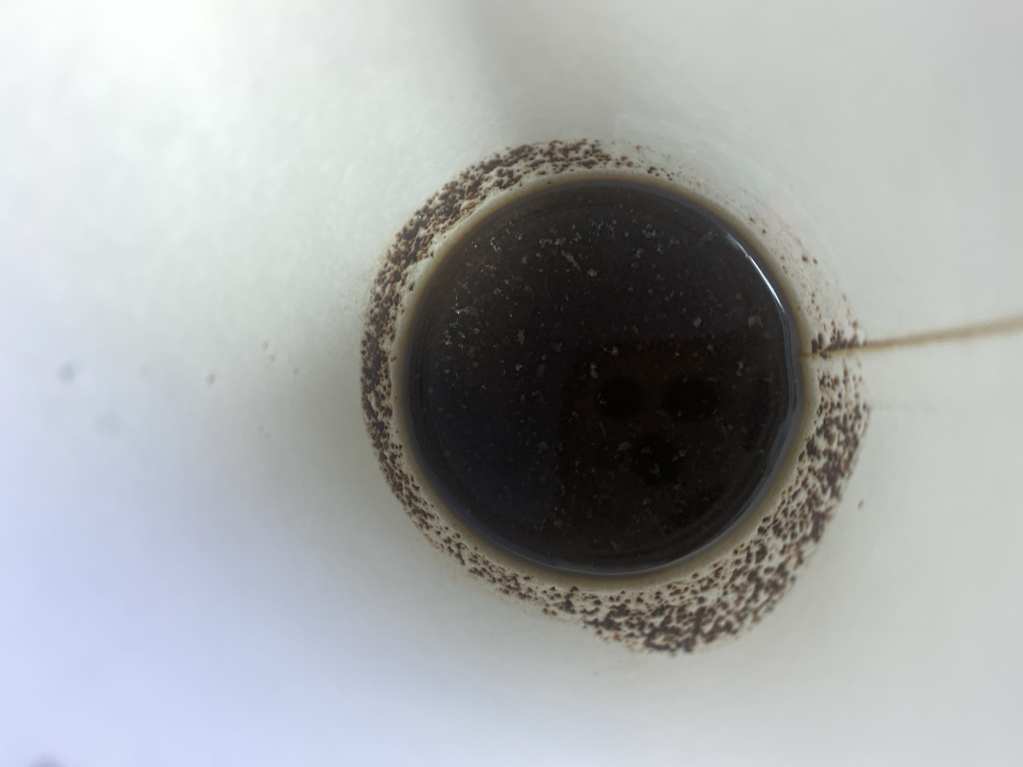 A top-down, close-up shot shows the inside of a white mug containing dark liquid, likely coffee. The liquid has some small particles suspended in it. A ring of brown residue, possibly coffee grounds, adheres to the inner rim of the mug. The mug appears to be made of ceramic or a similar material. The background is mostly out of focus and appears to be a light-colored surface. There are no people, animals, or other discernible objects present. The lighting suggests an indoor environment. This image does not provide any information that would allow for a determination of its location in Otaci, Moldova.