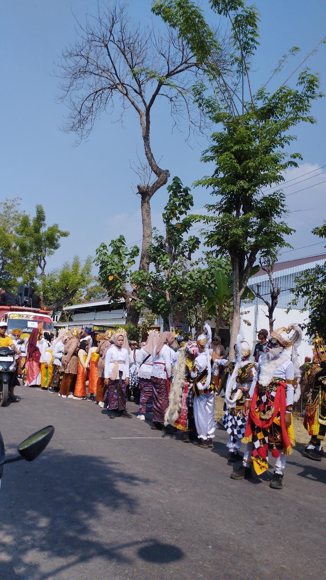 On a sunny day in the city of Bancar, Indonesia, a vibrant procession is underway. A group of people, dressed in traditional attire, are lining up on the asphalt road. Some are wearing white shirts and colorful sarongs, while others are adorned in elaborate costumes with masks and headdresses. The procession appears to be part of a cultural event or celebration. In the background, trees and buildings form the urban landscape. The presence of a motorcycle and a red truck suggests that this event is taking place in a public area. The scene is filled with color and movement, capturing the essence of Indonesian culture.