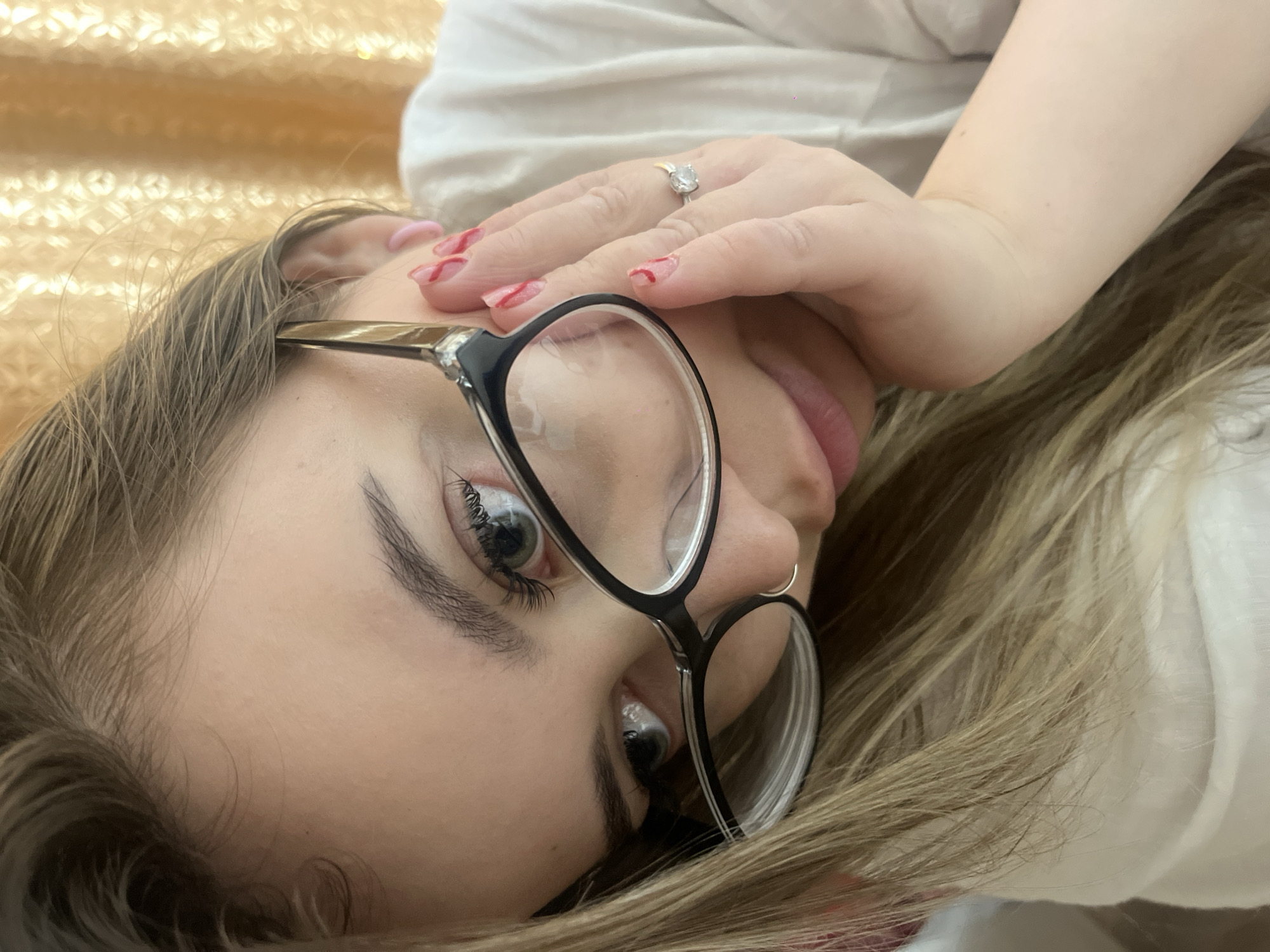 A person with light brown hair and glasses is looking directly at the camera from a reclined angle. The person is wearing a white shirt. The person's left hand is near their face, and a ring is visible on the fourth finger. A small hoop earring is visible in the person's nose. A gold-colored, textured background is visible. The location is Novopokrovskaya, Russia.