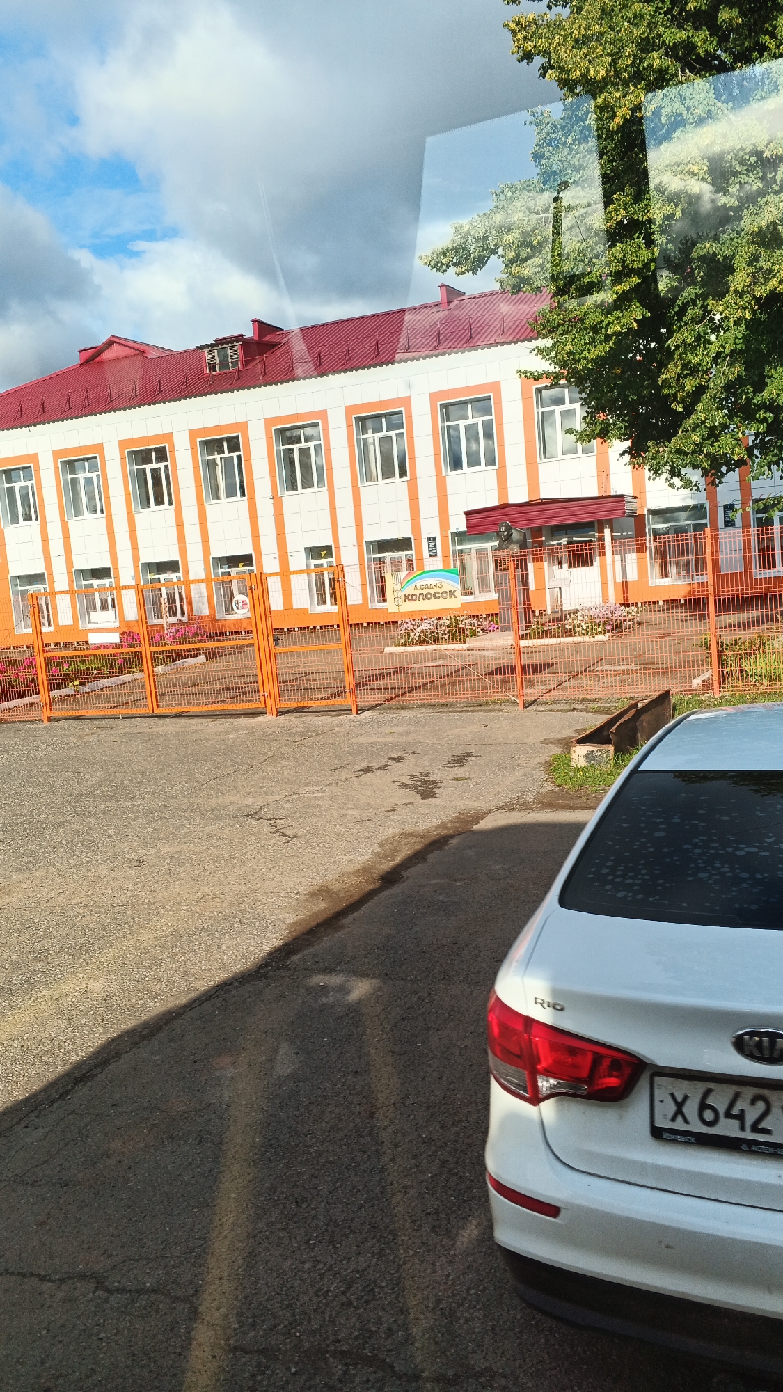 A white Kia Rio car is parked on an asphalt road in front of a two-story building with a red roof and orange and white exterior. The building appears to be a school or kindergarten, with a sign in Russian that reads "Детский сад КОЛОСОК" which translates to "Kindergarten Kolosok". There is an orange metal fence in front of the building with flowers planted behind it. The sky is partly cloudy. The license plate of the car is partially visible and reads "X642". The image is taken from a slightly elevated angle, likely from a vehicle.