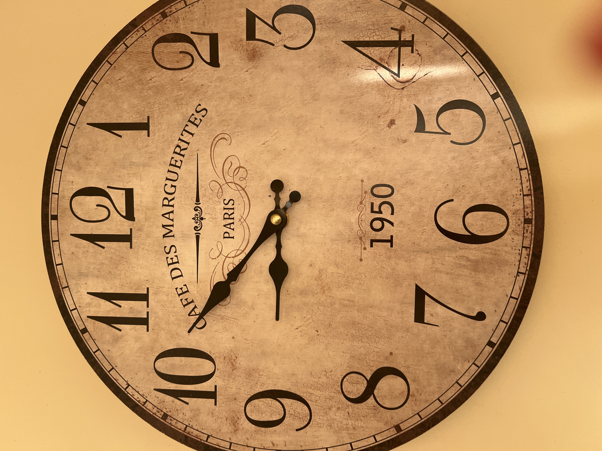 This is a close-up, eye-level, medium shot of a round, vintage-style wall clock. The clock face is a beige, textured material with Arabic numerals indicating the hours from 1 to 12. The clock has two black hands, with the hour hand pointing slightly past the 10 and the minute hand pointing to approximately the 11. The clock face also features decorative text that reads "CAFE DES MARGUERITES PARIS" and the year "1950" prominently displayed below the center. The clock is mounted on a plain, light yellow wall. The image does not contain any information that would allow for the identification of its location in Minsk, Belarus, or any details about people, actions, or interactions.