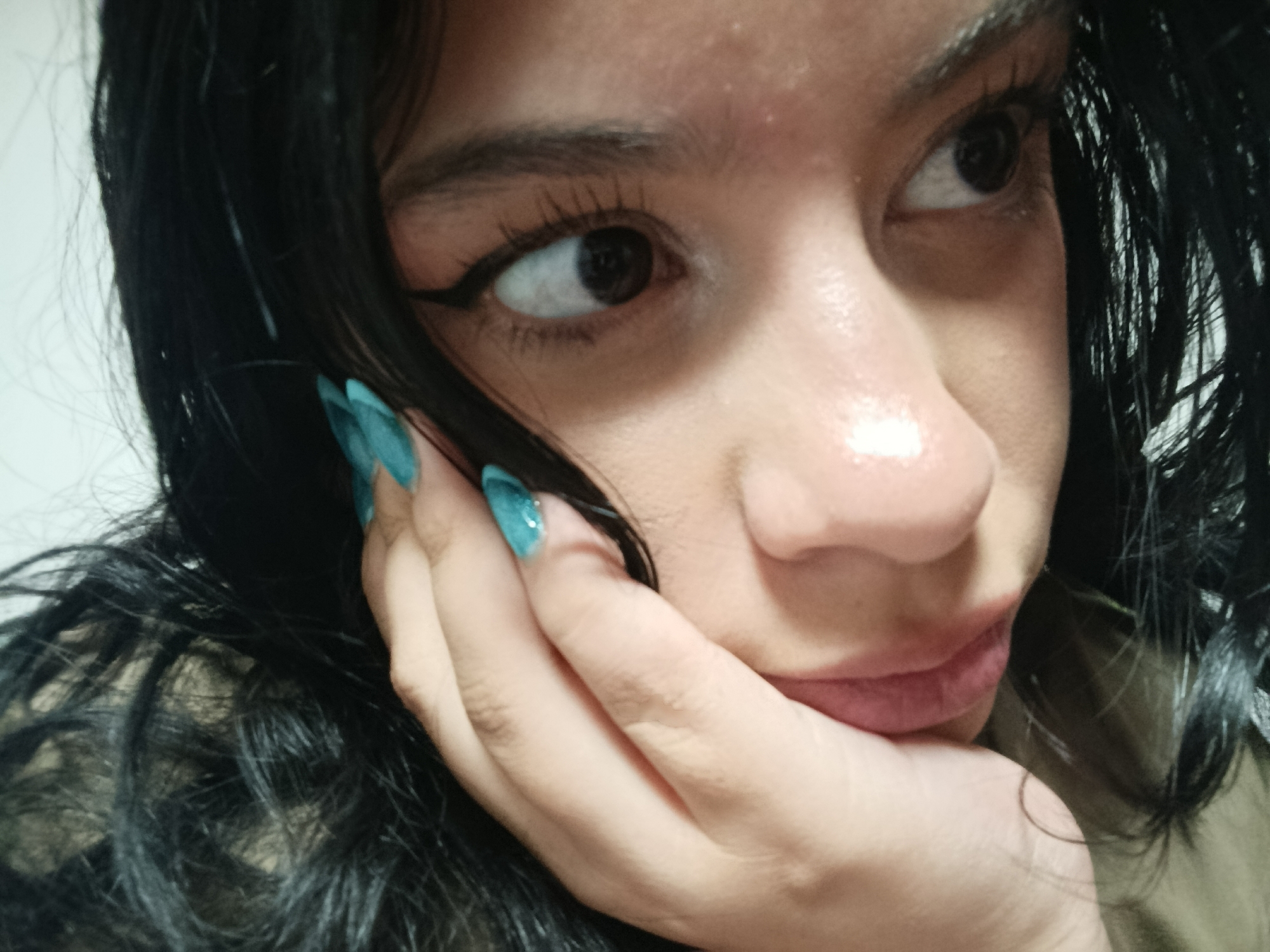 A close-up, eye-level portrait captures a person's upper face, dark hair, and hand. The subject, likely female, has dark, well-defined eyebrows, prominent black winged eyeliner, and long eyelashes, indicating makeup application. Their dark eyes gaze towards the right side of the frame, contributing to a thoughtful or observant expression.
The person's skin appears smooth, with a subtle sheen or highlight visible on the bridge of the nose and forehead. Part of their mouth and chin are visible, with naturally pink or subtly colored lips. Dark, possibly black, hair frames the left side of the face and falls around the neck area, appearing thick and slightly wavy or curly.
A hand is positioned with fingers resting against the chin and lower cheek, supporting the face. The fingernails are painted a distinctive turquoise or teal color, possibly with glitter or a textured finish, and have a pointed or almond shape. The background is uniformly light-colored and out of focus, suggesting a plain wall. There are no discernible objects beyond the person and their features, nor any visible text or specific location cues within the image itself. The lighting is soft and appears to be internal or indirect.
