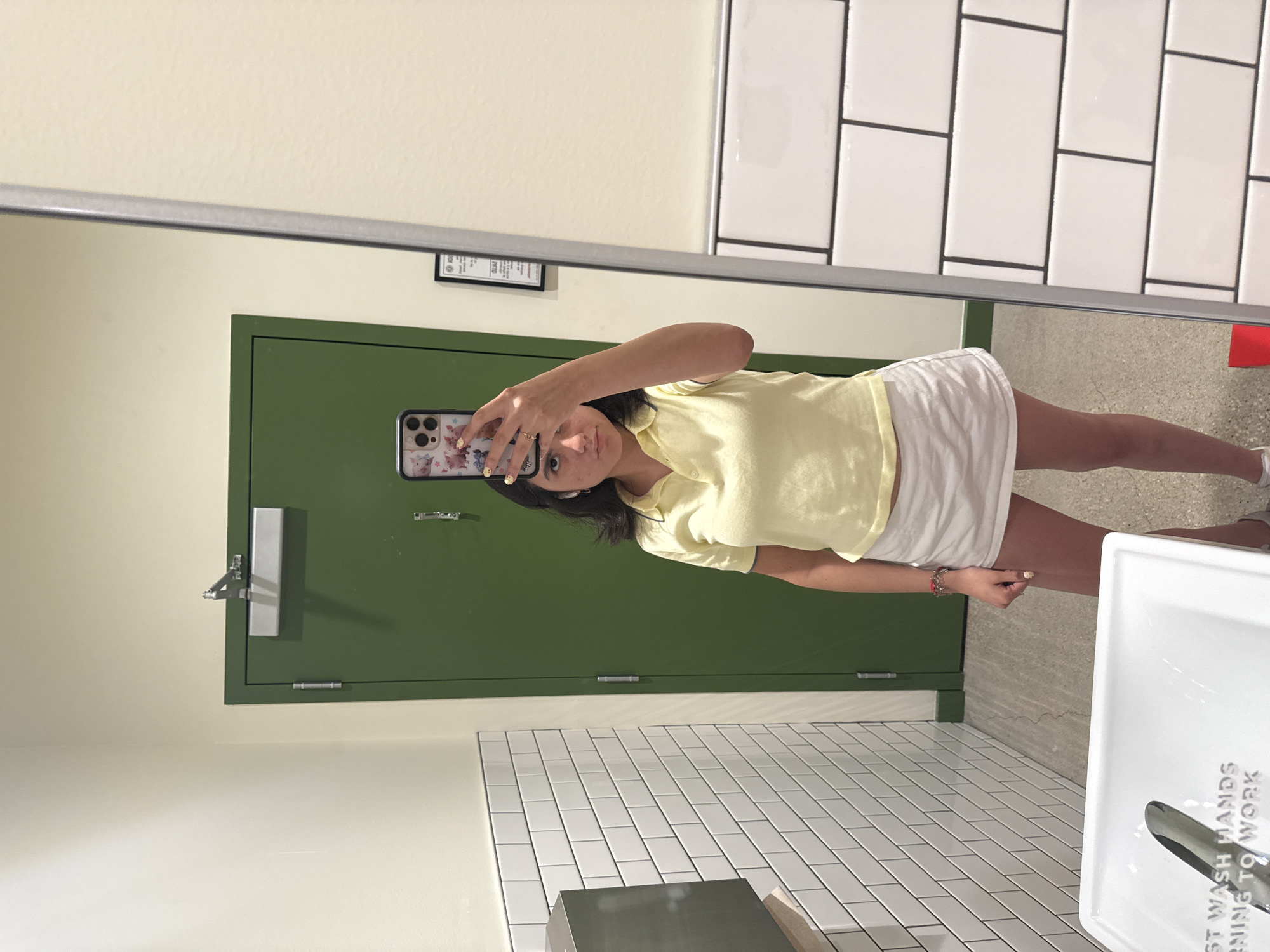 A young woman is taking a mirror selfie in a restroom. The restroom features white subway tile walls with black grout, a green door with a silver handle and hinges, and a white sink with instructions to wash hands before work. The sink has a chrome faucet. The woman is wearing a yellow collared shirt and a white skirt. Her phone has a decorative case. The location is likely in a business within Austin, Texas, given the typical style of restroom fixtures in public spaces.