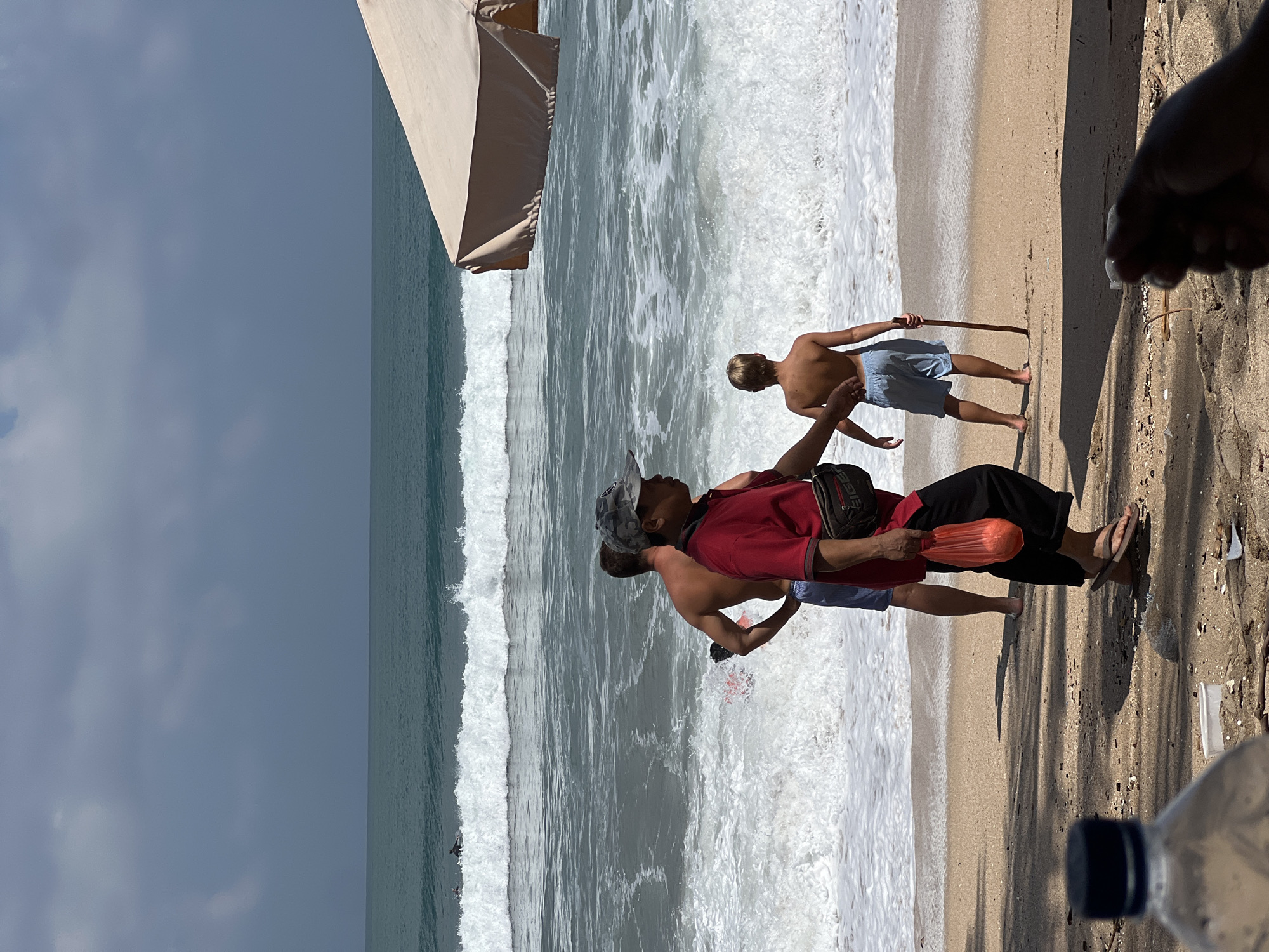 The image depicts a sunny day on Kuta Beach, Bali, Indonesia, with the ocean waves crashing onto the sandy shore. In the foreground, a person wearing a red shirt and black pants, holding an orange plastic bag and wearing flip-flops, walks towards the right of the frame. Behind them, a shirtless man in blue shorts stands with his hands on his hips, facing the ocean. To his right, a young boy in blue shorts walks towards the water, holding a wooden stick. In the background, surfers can be seen in the turquoise ocean, and a large beige beach umbrella is partially visible on the right. The sand is scattered with debris, including a plastic bottle in the lower left corner. The sky is partly cloudy, with a mix of blue sky and gray clouds.