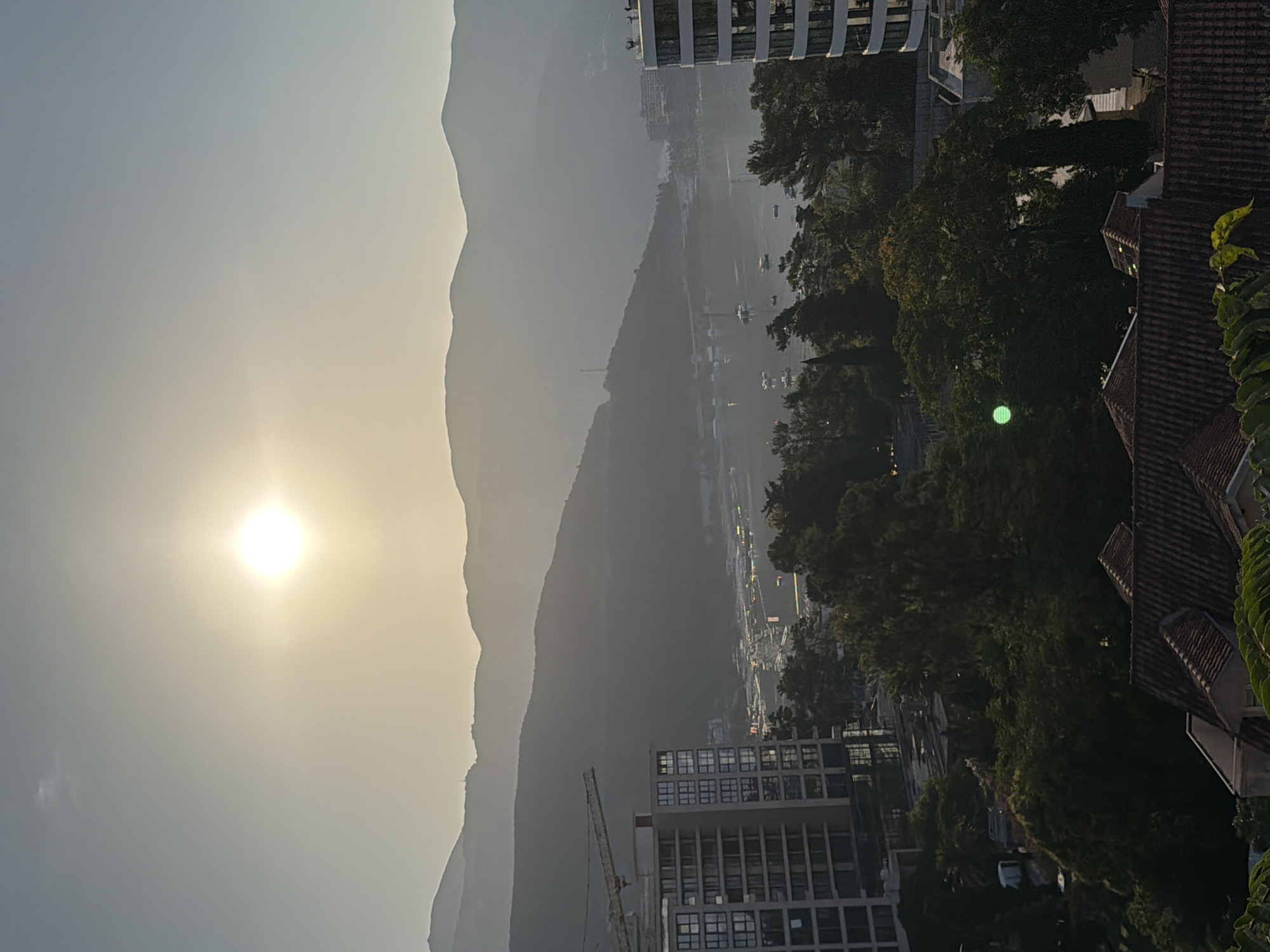 The image depicts a hazy, sun-drenched aerial view of a coastal city, likely Budva, Montenegro, during daylight hours. The sun, a bright orb, is positioned in the upper center of the frame, casting a soft glow and contributing to the atmospheric haze that softens the distant mountain ranges forming the backdrop. In the foreground, modern buildings with distinct architectural features, including balconies and large windows, are visible. One building on the left appears to be under construction, indicated by a prominent yellow crane. A body of water, possibly a bay or harbor, lies between the buildings and the mountains, dotted with numerous small boats and yachts. Dense green foliage, consisting of trees and other vegetation, covers parts of the landscape, adding a natural element to the urban scene. The scene is one of a developed coastal area, likely a resort or residential district, with a blend of natural beauty and constructed infrastructure.