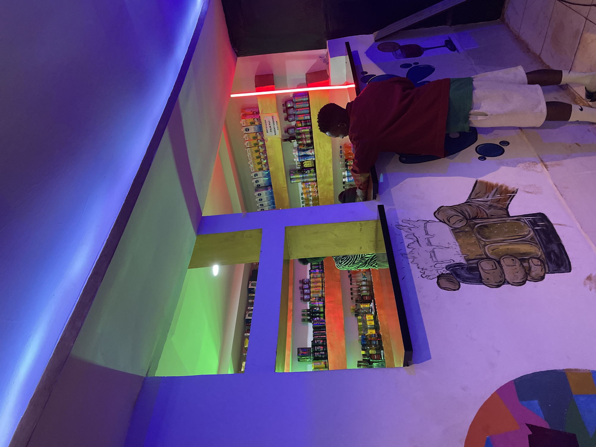 The image depicts the interior of a bar in Durumi, Nigeria, illuminated by colorful LED lights. In the foreground, a young man wearing a red hoodie, a green t-shirt, and white shorts stands with his back to the camera, reaching for items on a shelf behind a counter. The counter is decorated with a mural of a hand holding a frothy beer mug and abstract shapes. Behind him, shelves are stocked with various beverages, including bottles and cans. Above the counter, more shelves are visible, displaying more drinks and some boxes. The walls are painted in vibrant colors, and the ceiling is adorned with blue and green LED strip lights, creating a lively atmosphere. A portion of a person in a zebra-print shirt is visible in the background through an opening in the shelves.