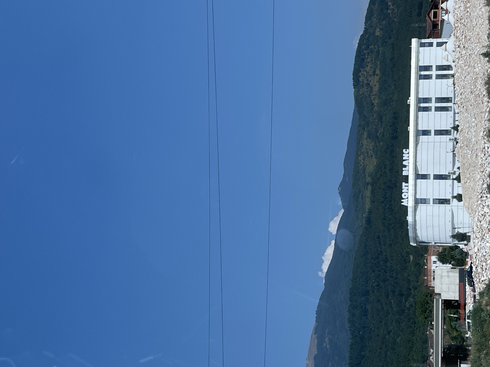 In Zdunje, North Macedonia, a modern, multi-story white building with prominent "MONT BLANC" signage stands under a clear blue sky. The building has a distinct architectural style with numerous rectangular windows. In front of the building, a pile of white rocks forms a foreground. Behind the structure, a densely forested mountainside rises, with a lighter, possibly cloud-dusted peak in the distance. Power lines are visible across the sky. To the left of the Mont Blanc building, there are other structures, including what appears to be a bridge or overpass under construction, with concrete and metal elements. A few vehicles are parked near the construction area. The overall scene suggests a developing area or a roadside commercial establishment in a mountainous region.