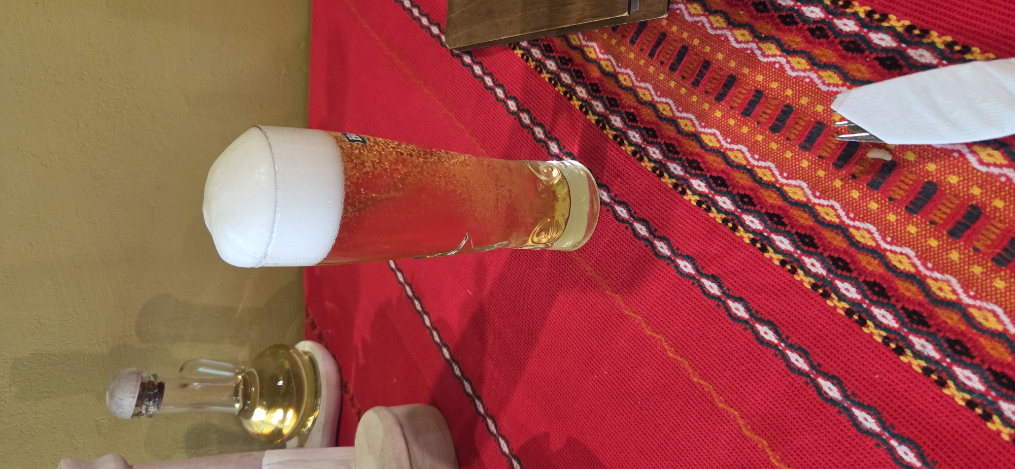 The image depicts a static tabletop scene, likely within an establishment in Beli Osam, Bulgaria. A tall, clear glass, approximately two-thirds full of a golden-amber liquid consistent with beer, occupies the central-right portion of the frame. Numerous small effervescent bubbles are visible rising within the liquid. A substantial, dense, white foam head, slightly mounded, caps the beer and protrudes slightly over the rim of the glass on the left side. Condensation is present on the upper exterior surface of the glass. A faint blue and white logo or text, possibly "Heineken," is visible on the glass's side.
The table is draped with a vibrant red woven tablecloth featuring traditional patterns. A diagonal band of repeating white, black, and thin yellow/orange geometric motifs crosses the tablecloth from the lower left to the upper right. Towards the lower right, a wider section displays a denser pattern with a predominantly orange-yellow base and red, black, and white rectangular and zigzag elements.
On the upper left, a small glass cruet, topped with a light-colored wooden stopper, contains a clear, golden-yellow liquid, consistent with olive oil. This cruet rests on a small, round, light-colored wooden coaster. Adjacent to the cruet, a larger, partially visible light-colored wooden object, possibly a grinder, stands upright. Below these, a small, round, lidded wooden box is also partially visible.
In the lower right corner, a silver-colored metal fork is wrapped in a white paper napkin. Next to this, a narrow, dark-colored wooden object, possibly a menu or bill holder, lies flat on the tablecloth.
The background consists of a plain, textured wall painted in a muted, yellowish-ochre color. Shadows are cast upon the wall from the objects on the table, particularly behind the oil cruet. No individuals are present in the image.