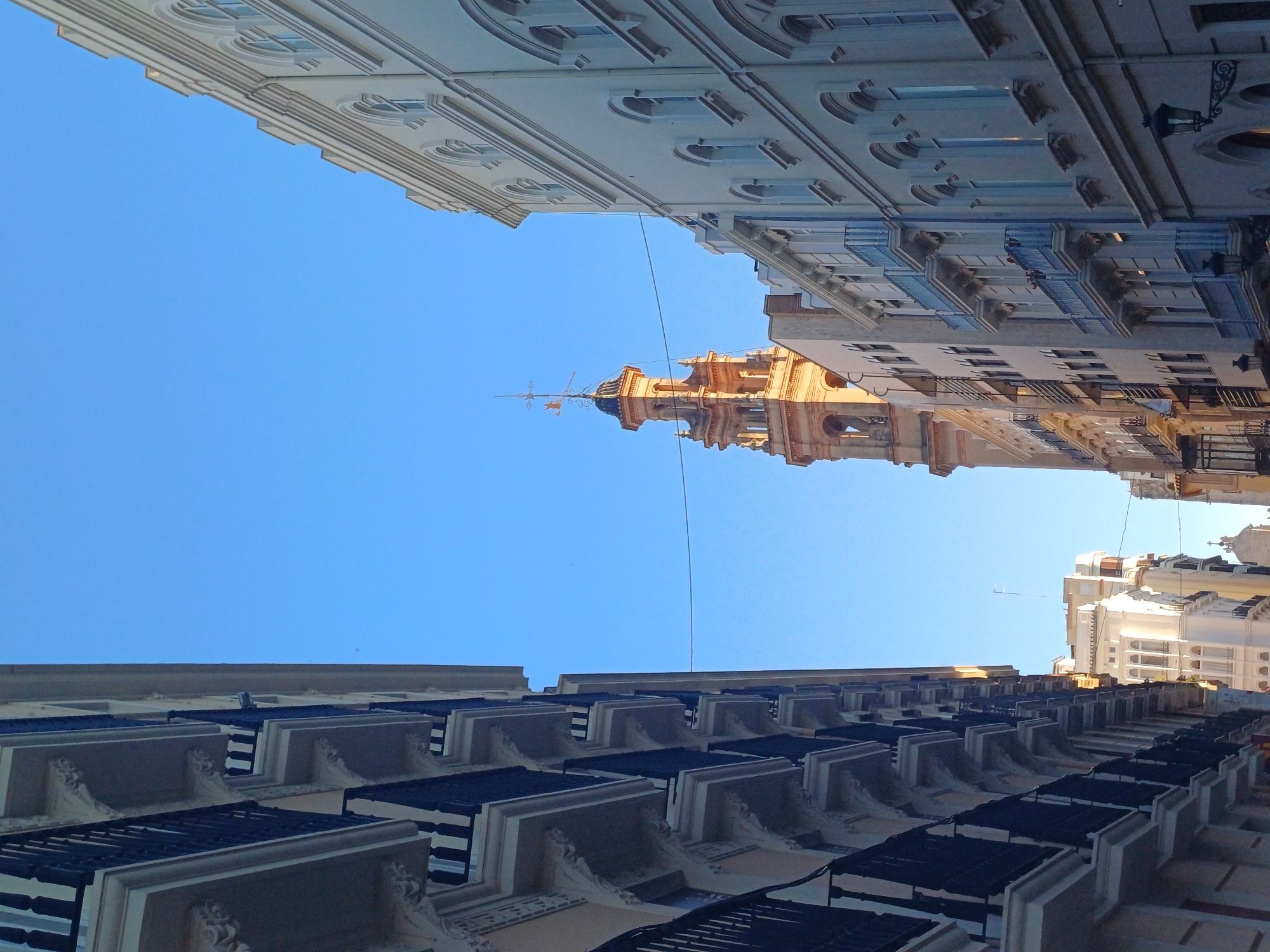 The image captures a street view in Valencia, Spain, taken from a low angle looking upwards. Architectural details of buildings line the street, with a clear blue sky visible between them. A notable tower structure is centrally positioned, receiving sunlight on its ornate facade. The buildings exhibit a range of styles, with visible balconies and architectural features. No people are directly visible in the shot. A wire extends across the sky between the buildings.
