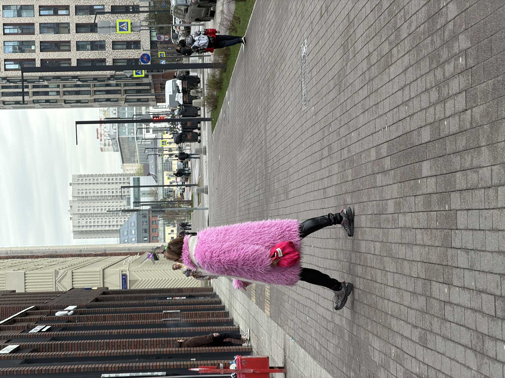The image, originally rotated 90 degrees clockwise, captures an urban street scene on an overcast day, likely in Moscow, Russia. The central subject is a person walking away from the viewer on a gray paved sidewalk, prominently wearing a vibrant, fluffy, bright pink faux-fur coat, black leggings, and dark sneakers. They are holding a small pink bag that complements their coat.
Several other pedestrians are visible in the background and mid-ground, engaged in walking. One person with a white backpack is seen further down the path, and another in a brown jacket stands near a stepped dark brick building in the foreground on the left. The street is lined with various modern buildings, including tall structures with light-colored, patterned facades and numerous windows, as well as darker brick buildings with distinct architectural textures.
Street infrastructure includes black lampposts, a red trash bin in the immediate foreground, and several dark gray dumpsters further along the path. Yellow square signs indicating a pedestrian crossing and a blue circular sign denoting a bicycle path are visible. Parked vehicles, mostly light-colored, can be seen in the distance. The sky is uniformly overcast, creating a soft, diffused light across the contemporary city environment.