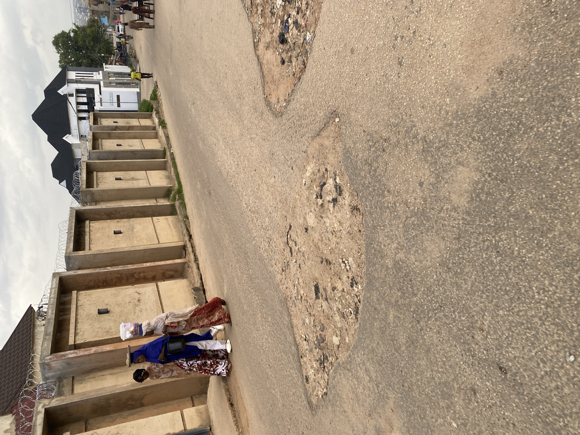 The image depicts a street scene in Jos, Nigeria. In the foreground, a paved road shows significant disrepair with large potholes. Along the left side of the road, a concrete wall topped with barbed wire separates the street from properties beyond. Three individuals are walking along the sidewalk adjacent to this wall. The first person, on the left, wears a patterned dress and a dark headscarf, carrying something on their head. The second person is in a blue outfit with white pants and a black bucket. The third person is wearing a light-colored headscarf and a long red and white patterned dress, also carrying items on their head. Further down the road, a modern building with a black, multi-peaked roof is visible on the right, with other buildings and trees in the background. Several other people and a motorcycle are present further in the distance, suggesting ongoing activity on the street. The sky is overcast.