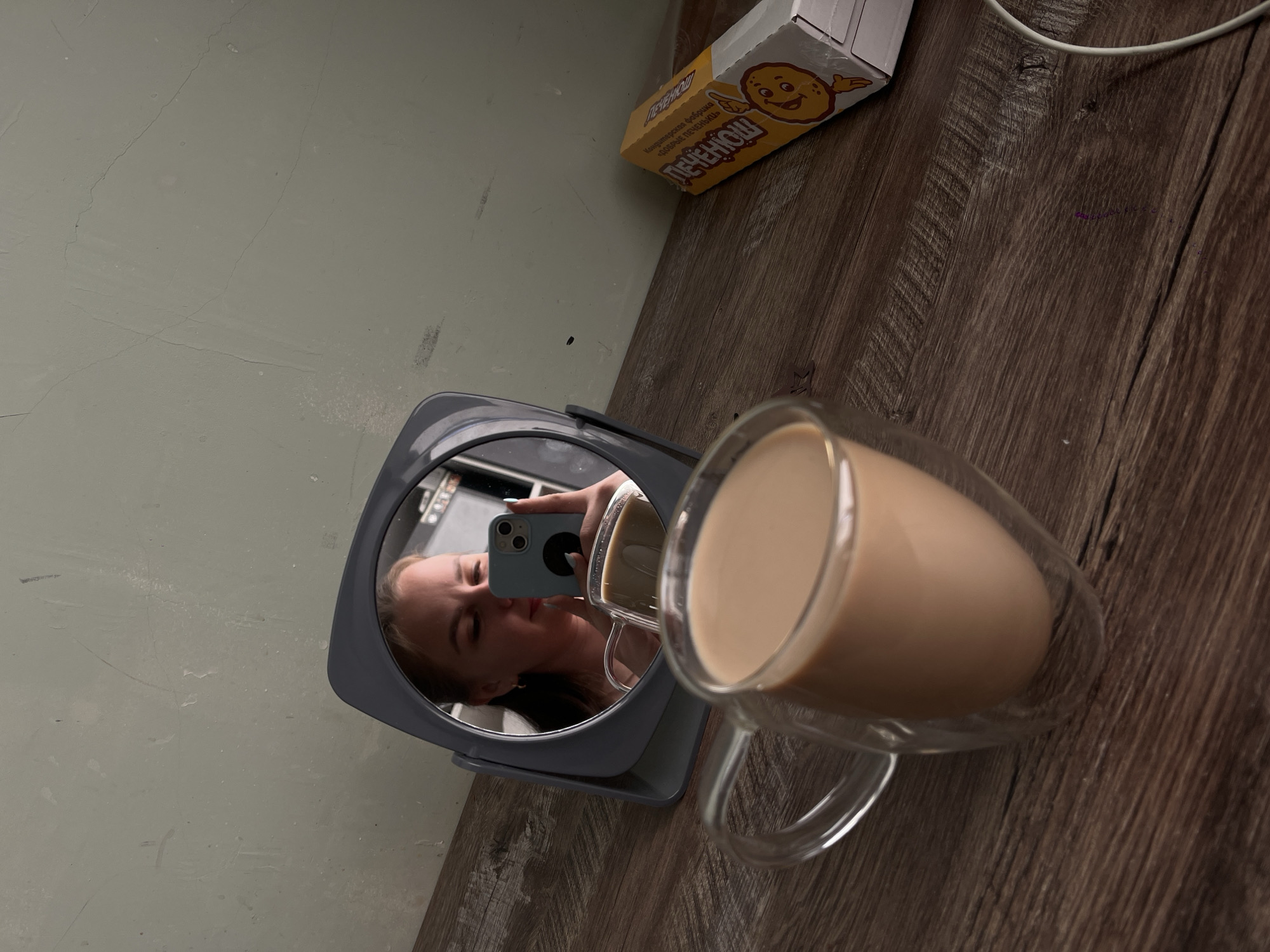 The image depicts a mirror reflecting a woman taking a photo of herself with a phone, with a mug of light brown liquid, possibly coffee or tea, in the foreground. Adjacent to the mirror is a package of "Pechenki" cookies. The background consists of a wall and a wood-patterned surface. The location in Izhevsk, Russia, is not discernible from the image.