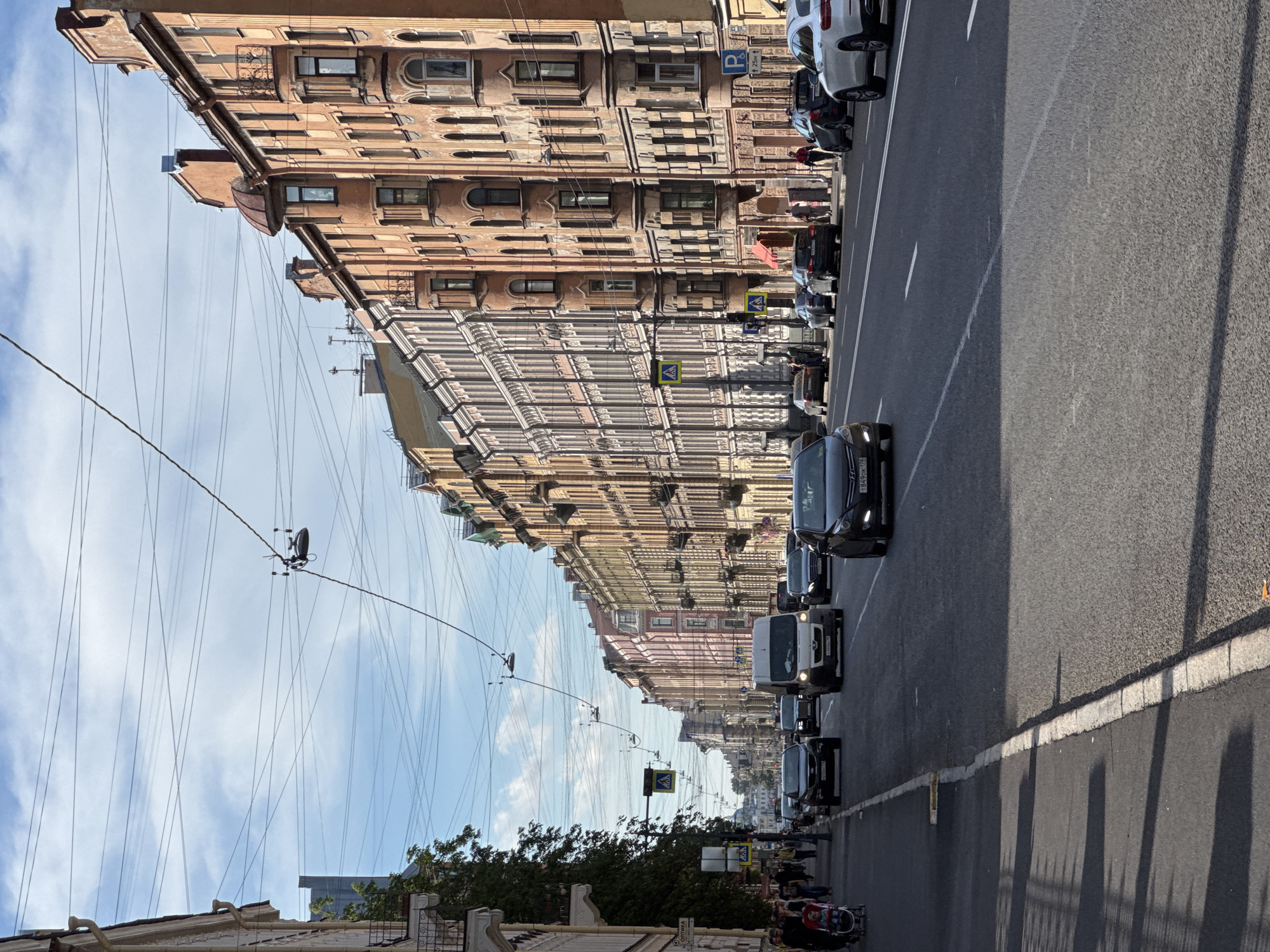 This image depicts a daytime view of a street in Saint Petersburg, Russia. The street is lined with multi-story buildings with ornate architectural details, characteristic of the city. Several vehicles, including cars and a van, are driving on the road. Pedestrians are visible on the sidewalk, some walking and one pushing a stroller. Overhead, a complex network of wires crisscrosses the sky, a common sight in many urban environments. The weather appears to be partly cloudy, with sunlight casting shadows on the street and sidewalk. The perspective is from street level, looking down the thoroughfare.
