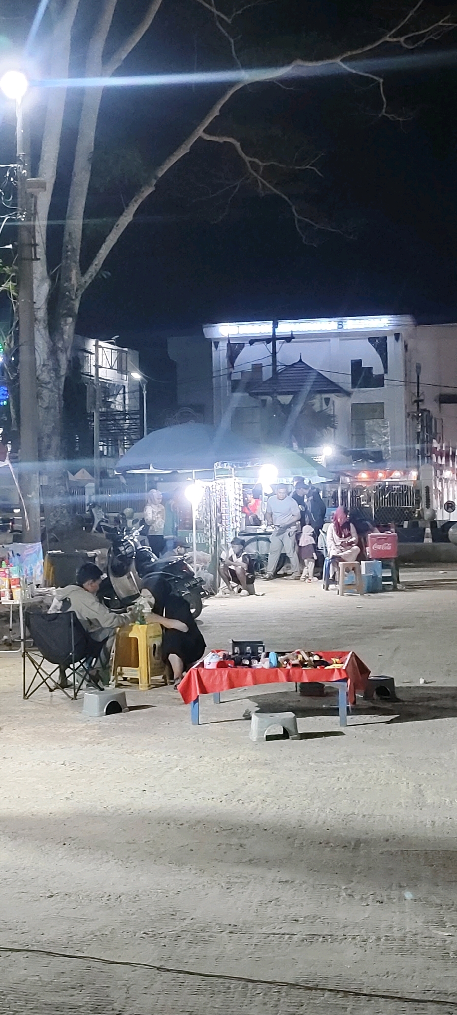 The image depicts a night scene at a market in Martapura, Indonesia, likely a temporary setup on a street or open area. In the foreground, a red-covered table displays various items, possibly merchandise, with several small plastic stools around it. Two individuals, a man and a woman wearing a hijab, are seated on chairs near a motorcycle, engaged in conversation or an activity.
Further into the scene, other individuals are visible, some standing and some seated. A woman in a hijab is sitting on a stool, and a young child stands nearby. Another group of people are gathered around a stall, with a Coca-Cola cooler visible. In the background, there are buildings with illuminated windows and awnings. Streetlights, including one prominent light source in the upper left, illuminate the area, casting shadows and creating a somewhat grainy and low-light atmosphere. The ground appears to be a dusty or sandy surface. The overall impression is of a bustling night market environment.