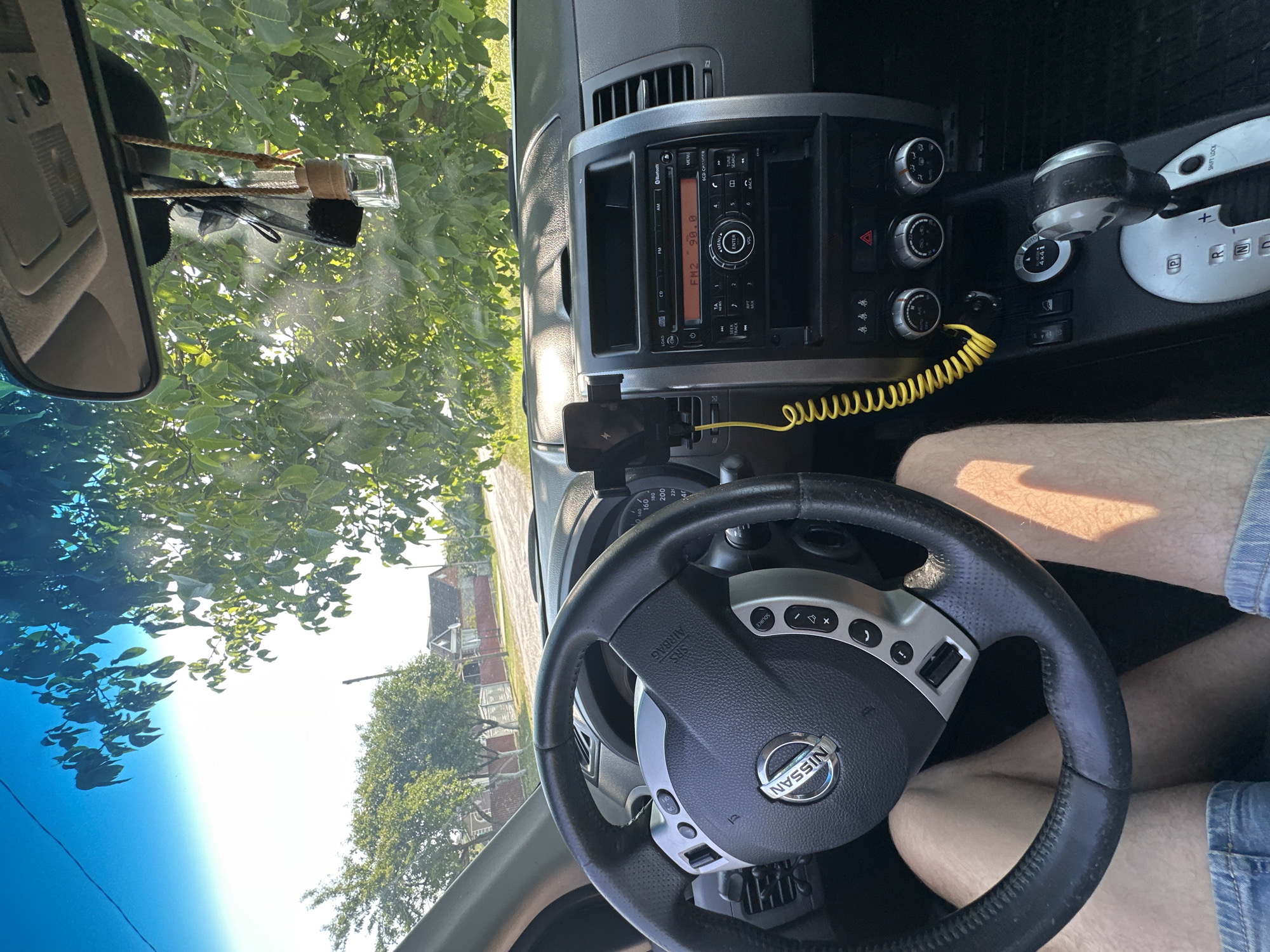 The image is a point-of-view shot from inside a car, looking forward. The car is a Nissan, as indicated by the logo on the steering wheel. The dashboard is visible, including the car stereo and climate controls. A smartphone is mounted on the dashboard, connected by a yellow cable. Outside the car, trees and houses are visible, suggesting a rural or suburban setting. The sunlight indicates it is daytime. The location is identified as Shatava, Ukraine.