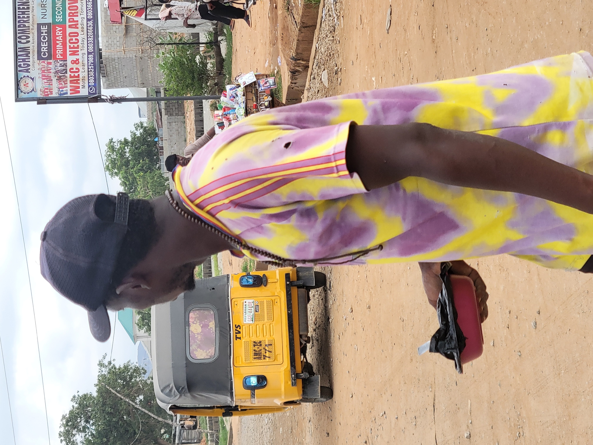 The image shows a man wearing a black cap and a yellow and purple tie-dye shirt, standing next to a yellow tricycle (TVS, registration GGE-444OR, Lagos). He is holding a small red object with black material protruding from it. In the background, there's a sign for "AGHAM COMPREHENSIVE," advertising Creche, Nursery, Primary, and Secondary levels, as well as WAEC & NECO approvals, with the motto "Learning Today, Leading Tomorrow," and phone numbers. The location is identified by the sign as being in Rafinsanyi New Layout, Kwamba, Nigeria. The ground is dirt or sand.