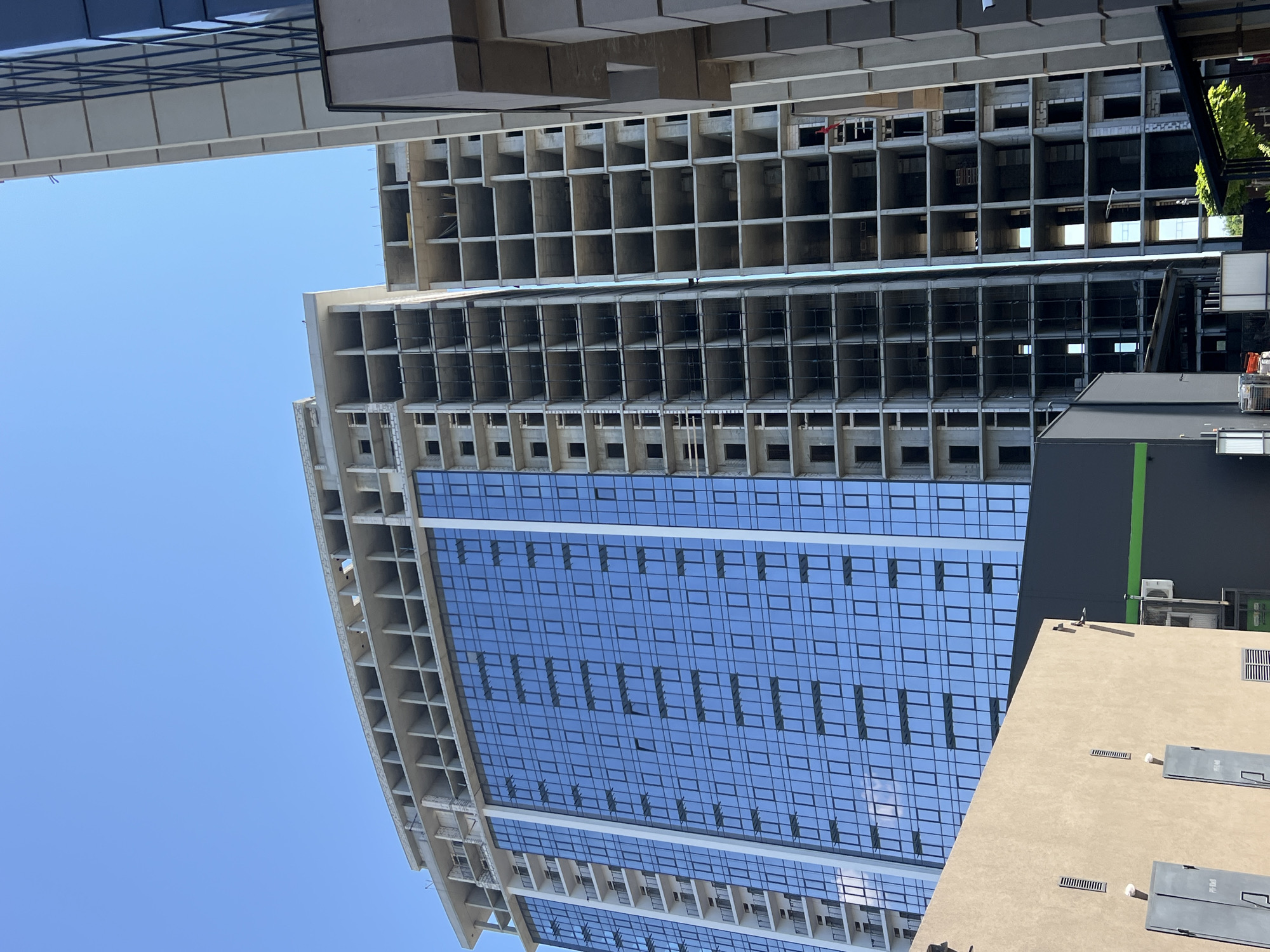 In Fontanka, Ukraine, under a clear blue sky, a partially constructed high-rise building dominates the scene. The left side of the building features a façade of blue glass panels, reflecting the sky and surrounding structures. The right side of the building is a concrete skeleton, exposing numerous unfinished rooms and balconies. Adjacent to this main structure, another, more complete building with a similar glass façade is partially visible on the far right. In the foreground, to the left, a low-rise beige building with grey doors and ventilation grates is visible. To the right of the beige building, a dark grey structure with a green horizontal stripe is situated. The construction site appears active, with construction elements visible on the unfinished building. The overall scene depicts an urban environment in development.