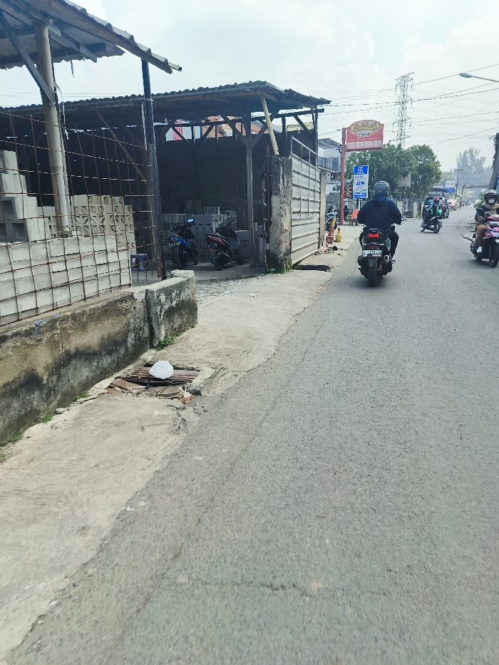 The scene is located in Cileunyi, Indonesia, at latitude -6.943 and longitude 107.726. It is a street lined with concrete blocks. A man on a motorcycle is driving down the street, away from the camera. There is a wooden building with a sign above it on the left side of the street. There is another motorcycle parked near the building, and two others parked further down the street. Behind the building there is a metal fence. There are trees and plants growing on the edge of the road. There are two other motorcycles parked on the side of the road, along with some other vehicles. The pavement is cracked and there is a white object lying on the ground. The sky is clear and the weather is sunny.