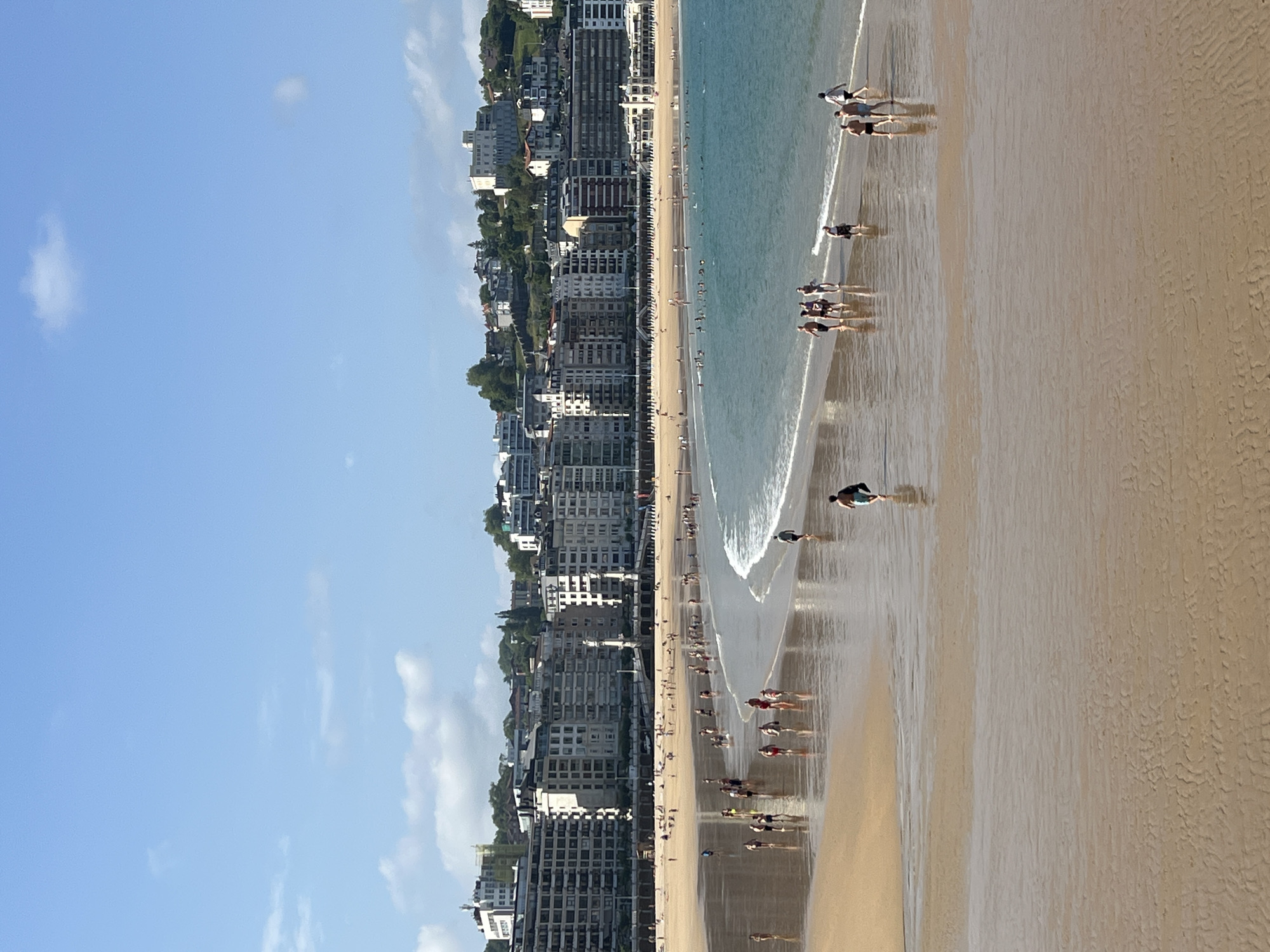 This image captures a wide view of La Concha beach in San Sebastián, Spain, on a sunny day. The foreground shows a wet, sandy beach with ripples from receding water, reflecting the light. Several individuals are scattered across the beach, some walking towards the water, others moving away. In the mid-ground, a gentle wave breaks along the shoreline, with more people visible wading in the shallows and swimming further out. The background features a crescent-shaped bay lined with a promenade and a dense collection of multi-story buildings, typical of the city's architecture. The buildings are a mix of cream, white, and beige, with many windows visible. Behind the buildings, a green, hilly landscape rises, dotted with more structures, and the sky above is a clear blue with a few wispy clouds. The overall scene conveys a sense of a bustling summer day at a popular urban beach.