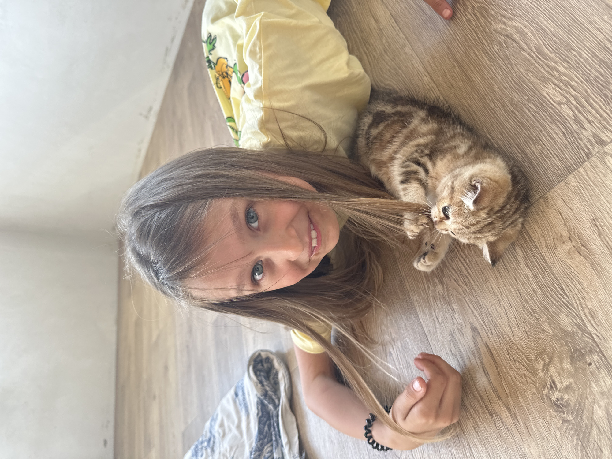 An over-the-shoulder medium shot shows a young girl with long, light brown hair and blue eyes, lying on a wooden floor and looking at the camera with a wide smile. She is wearing a yellow t-shirt with a pineapple print. A small, tabby kitten is lying next to her, on its back, with its paws in the air. The kitten is looking up at the girl, who is reaching out to pet it. The background is a plain white wall. The scene takes place indoors, on a wooden floor, likely in a home environment. It is not possible to determine the specific location in the city of Mokino, Russia from the image alone.