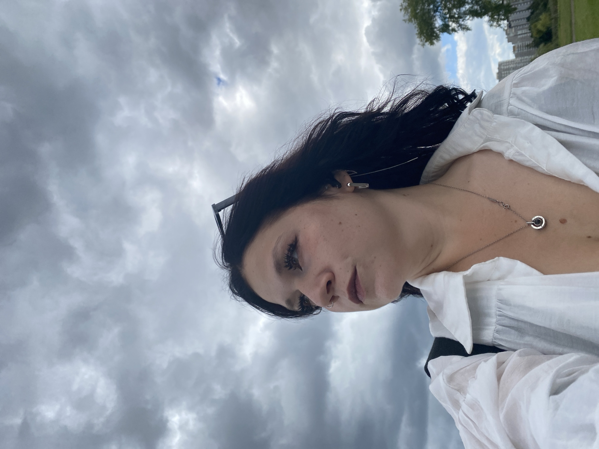 A medium shot taken from a low angle shows a young woman with dark hair and a nose ring looking to her left against a cloudy sky. She is wearing a white collared shirt with ruffled sleeves, a silver necklace with a circular pendant, and a rectangular earring in her left ear. Sunglasses are perched on top of her head. In the background, a city skyline with buildings and trees can be seen. The scene appears to be an outdoor urban environment.