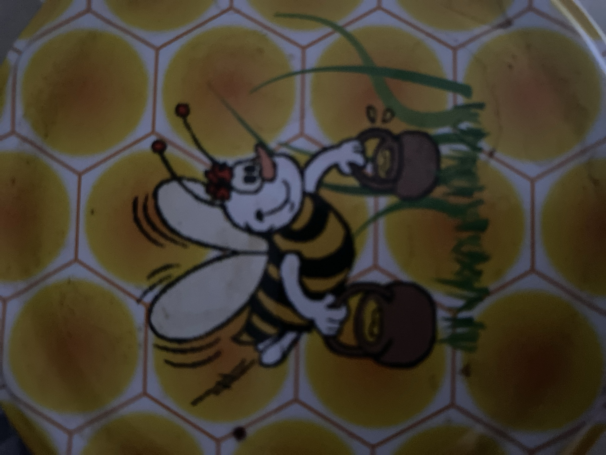 The image is a close-up of a cartoon drawing of a bee. The bee is white with black and yellow stripes and has large white wings. It is wearing a red flower on its head and has a smiling expression. The bee is holding two small brown baskets, one in each hand. The background is a honeycomb pattern with yellow hexagonal cells. There are also some green blades of grass depicted. The image appears to be from a tin or a similar decorative item. There is no discernible information in the image to indicate its location in Zgrade, Montenegro.