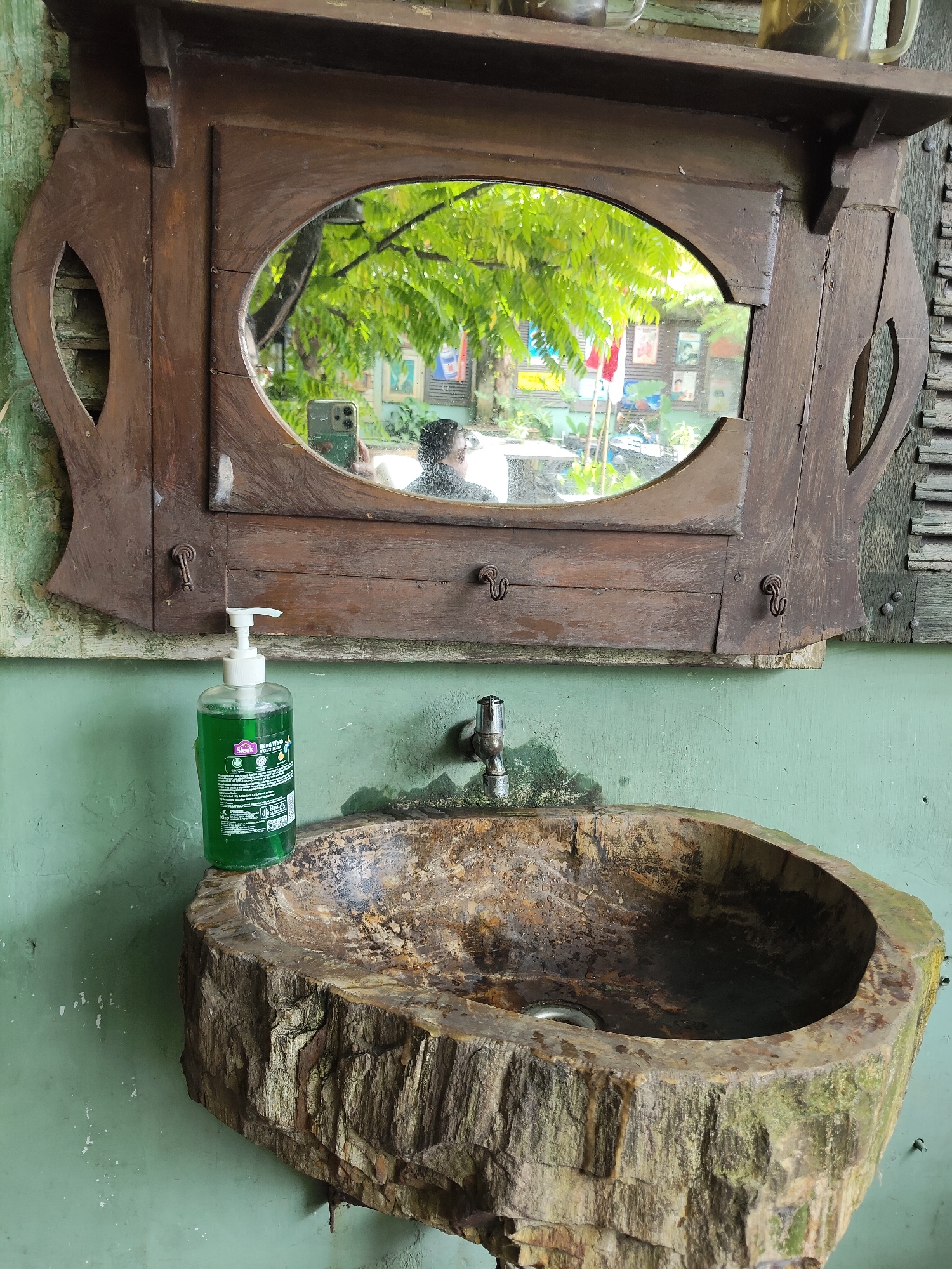 The image depicts an outdoor sink area, likely in the city of Medan, Indonesia, given the visual cues such as the greenery and what appears to be a tropical environment. The sink itself is a unique, rustic creation carved from a large piece of wood, with its rough exterior and hollowed-out basin showcasing natural textures. A silver faucet is mounted above the sink. To the left of the sink, a clear plastic bottle of green liquid hand soap with a white pump dispenser is prominently placed. Above the sink, a dark wooden mirror frame surrounds a mirror that reflects the environment. In the reflection, a person can be seen holding a mobile phone, suggesting they are taking the photograph. The background visible in the mirror shows lush green trees and foliage, indicating an outdoor setting. The wall behind the sink and mirror is painted a muted green color, showing signs of wear. The overall impression is one of a functional, yet artistic and environmentally integrated, wash basin.