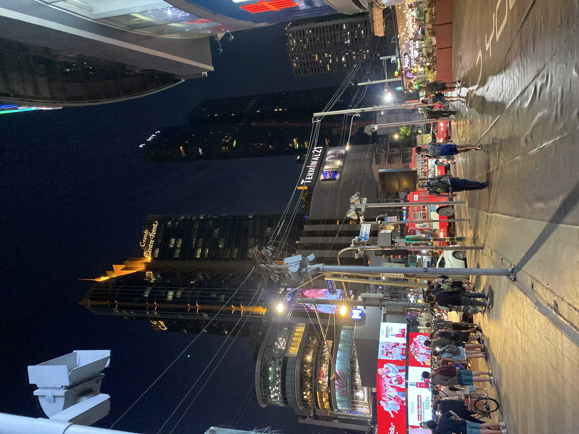 The image depicts a nighttime scene in Bangkok, Thailand, near the Terminal 21 shopping mall. Several tall buildings are visible, including the Grande Centre Point Hotel. Streetlights illuminate the sidewalk, where a crowd of people is gathered near a large electronic billboard advertising POND's skincare products. Across the street, more pedestrians are walking on the sidewalk. Double-decker tour buses can also be seen on the street. Utility poles with wires are present.