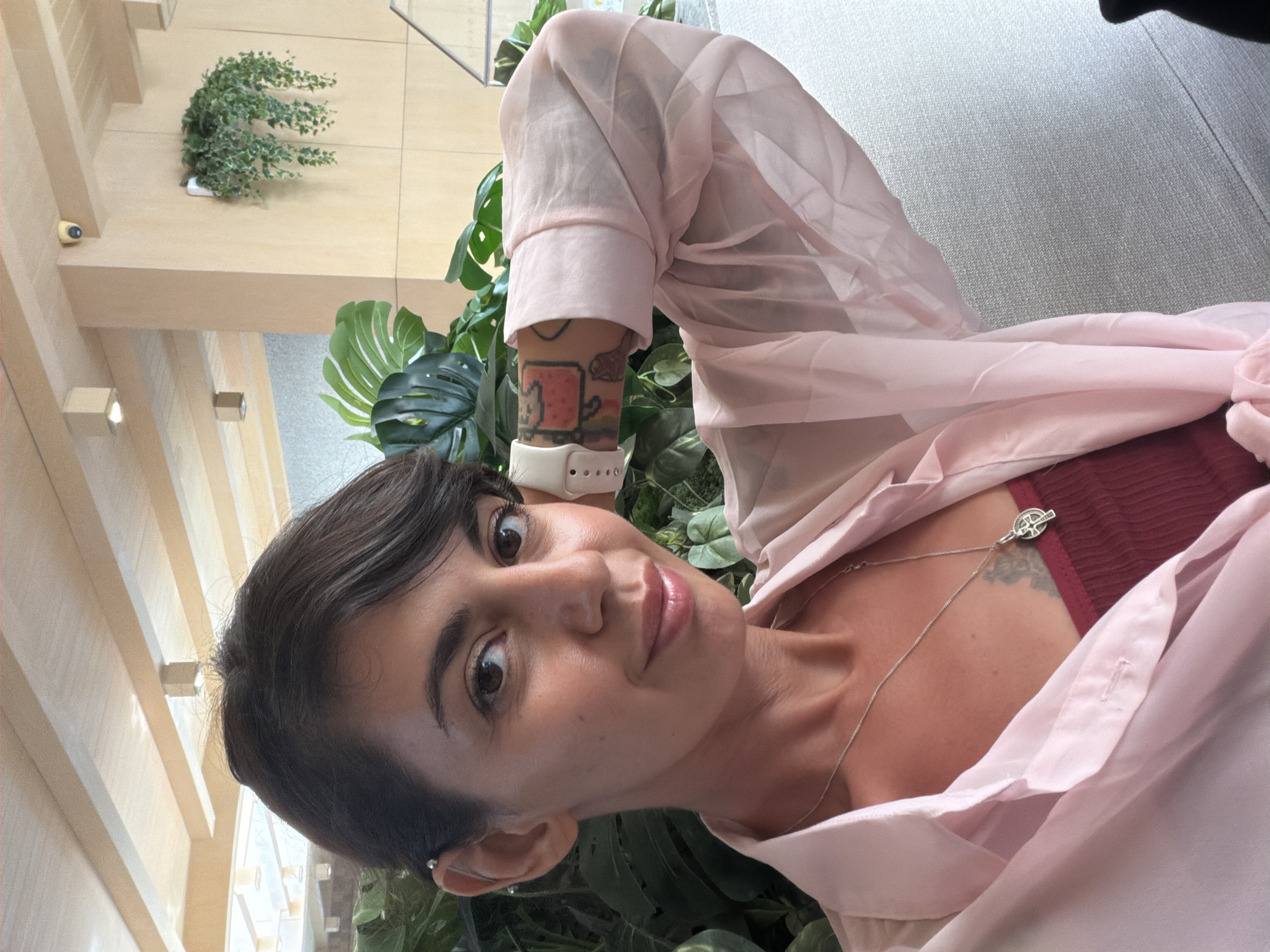 This image is a close-up, vertical portrait of a young adult taken indoors, likely in a modern building in North Kuta, Indonesia. The subject has short dark hair, brown eyes, and fair skin, looking directly at the camera with a subtle, pleasant smile.
She is wearing a sheer, light pink long-sleeved button-up shirt over a dark red ribbed tank top. A silver chain necklace with a small, intricate round medallion hangs around her neck. On her left wrist, she wears a white smartwatch, and a colorful tattoo of the Nyan Cat is visible on her left forearm. Another tattoo is partially seen on her upper chest/shoulder area. She has a small silver stud earring in her left ear.
The background features lush green indoor plants, including large Monstera leaves and a hanging vine. Light-colored wooden beams or slatted panels, along with light gray walls, indicate a contemporary interior design. High on one of the beams, a small, yellow-lensed security camera is mounted. The lighting suggests it is daytime. The subject appears to be posing for a casual portrait or selfie.