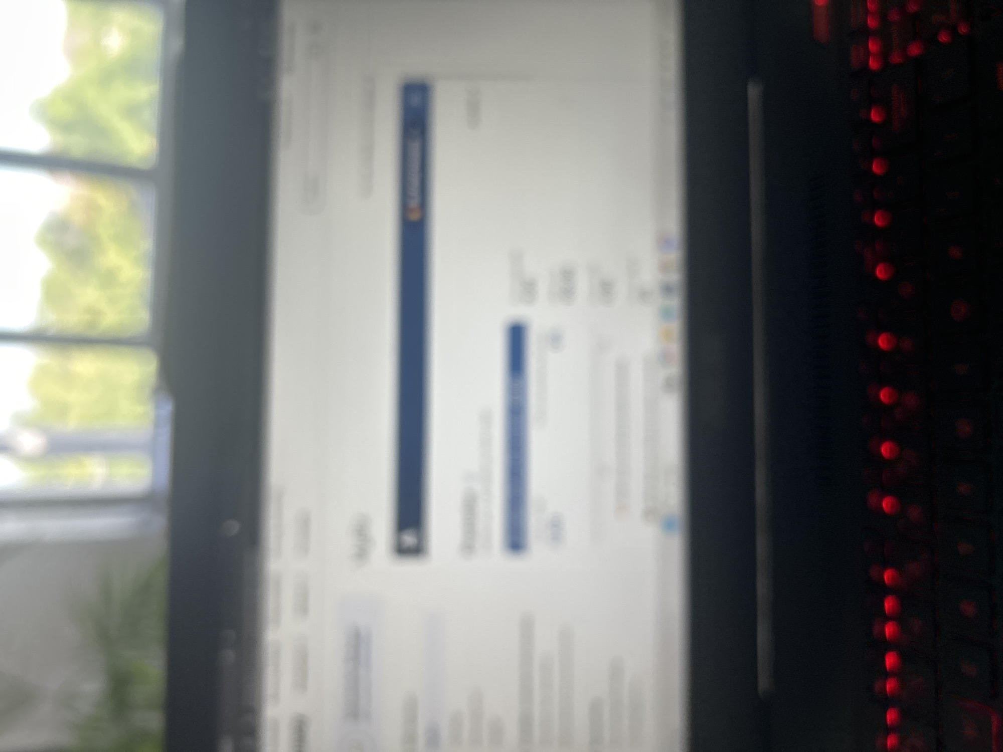 The image shows a close-up view of a laptop screen displaying a financial or accounting interface. The screen is somewhat blurred, but elements like columns with numerical data and headings are visible. The laptop itself has a dark keyboard with red backlighting, suggesting it is a gaming or performance-oriented model. In the background, out of focus, is a window with a view of greenery and trees, indicative of an outdoor environment. The scene appears to be set indoors, likely in a home or office setting, in Puurs, Belgium. There are no people visible in the image.